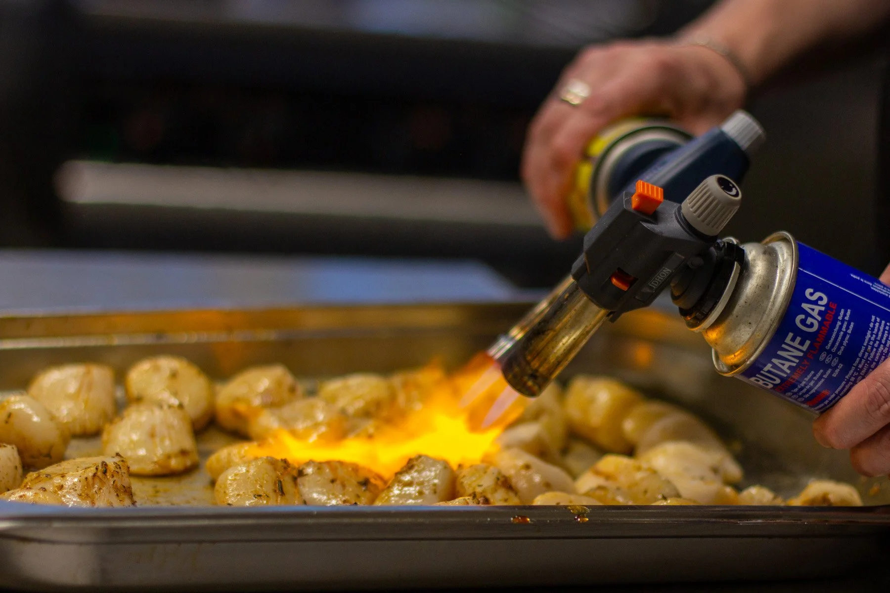 Someone using a culinary blowtorch to caramelize or brown food on a baking sheet. The person's hand is holding a butane gas canister attached to the torch.
