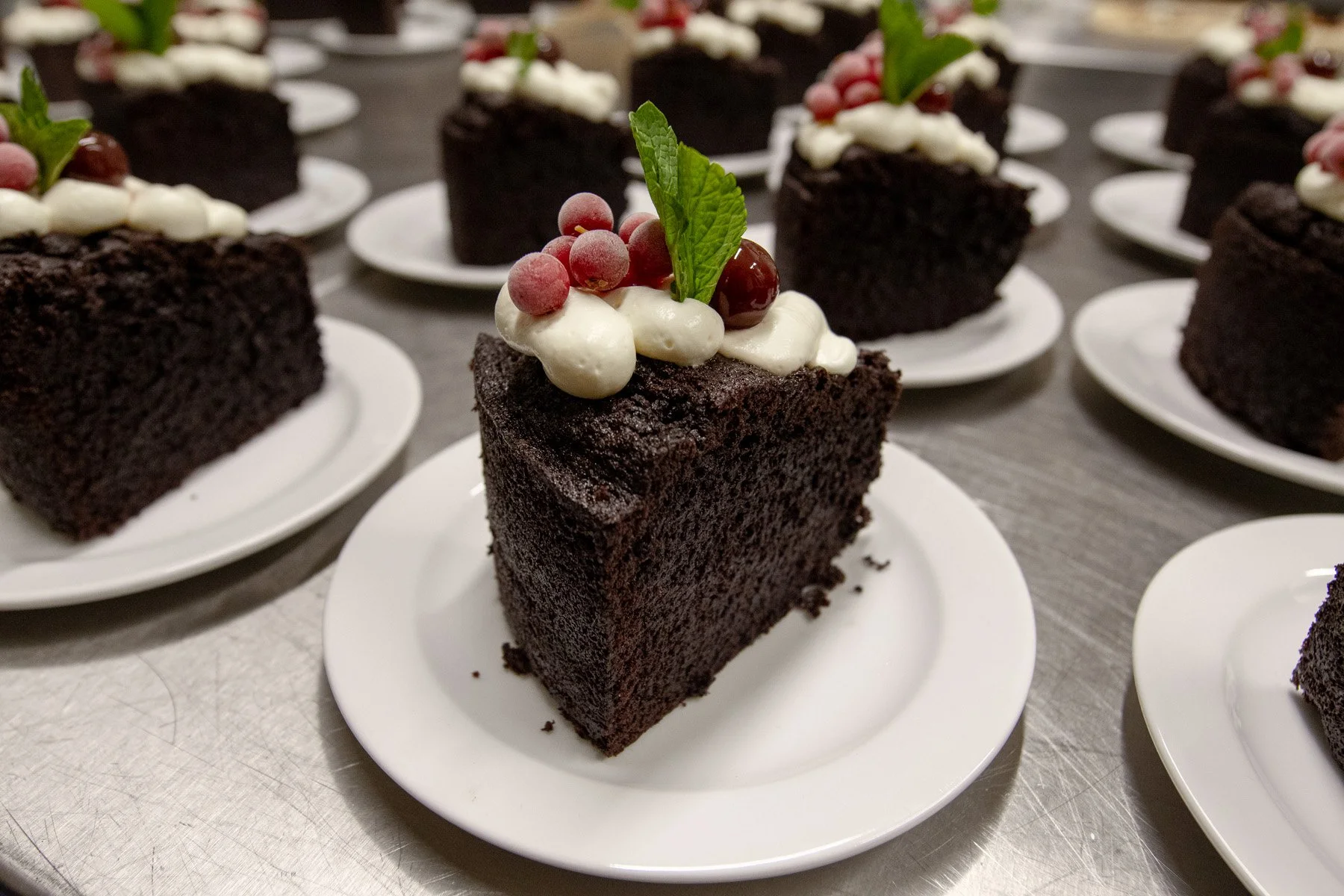 A slice of chocolate cake topped with whipped cream, red berries, a green mint leaf, and other small garnishes on a white plate, with several similar slices in the background.