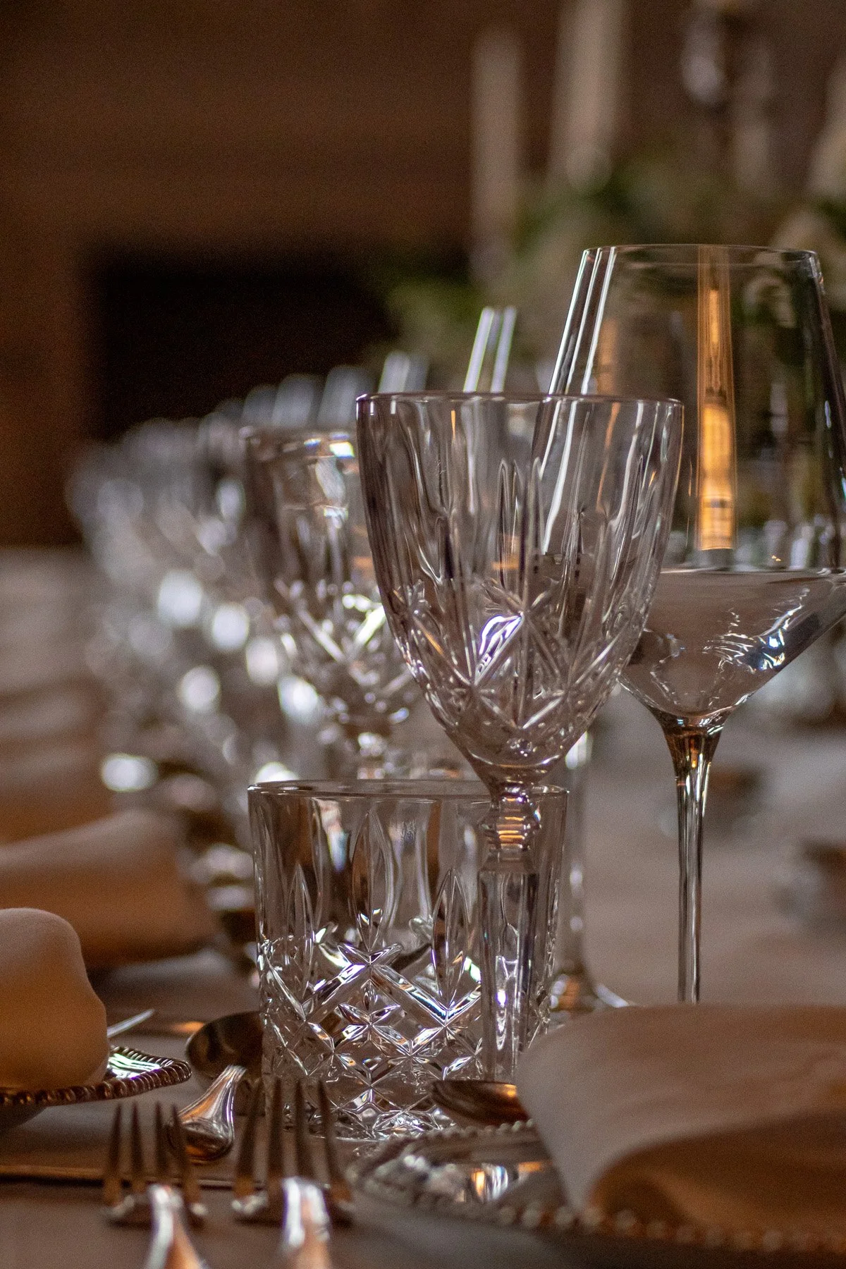 A close-up of a dining table setting with various empty crystal glasses, silverware, and a white napkin in the foreground.