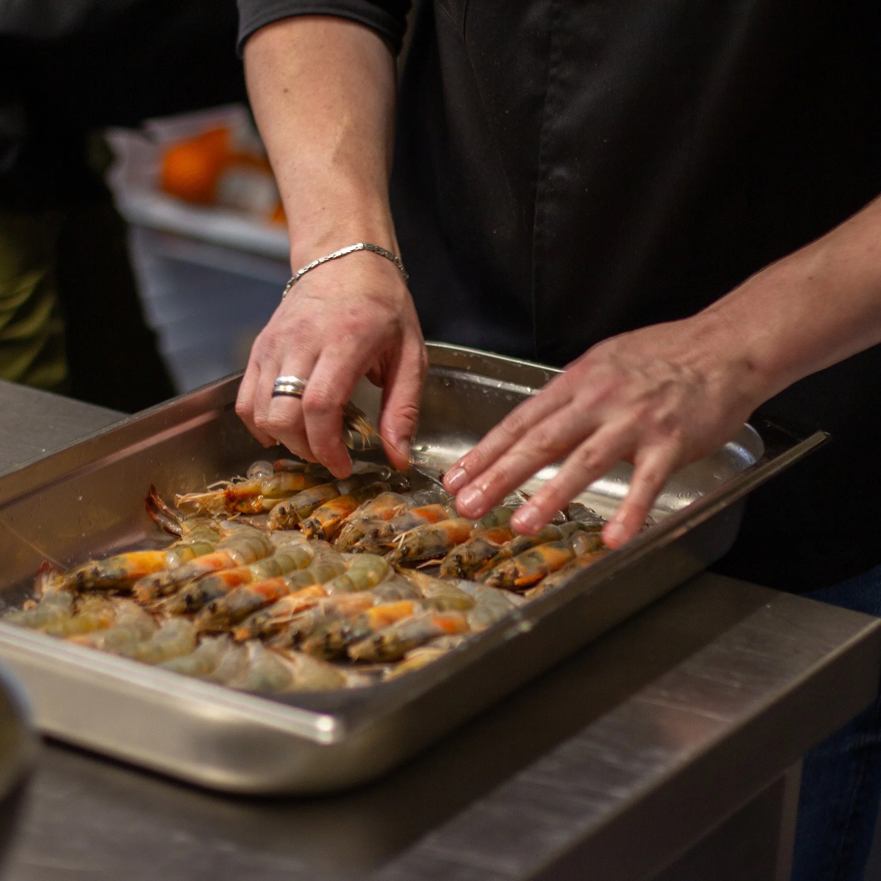A person preparing raw shrimp on a metal tray, arranging them with their hands in a kitchen or culinary setting.