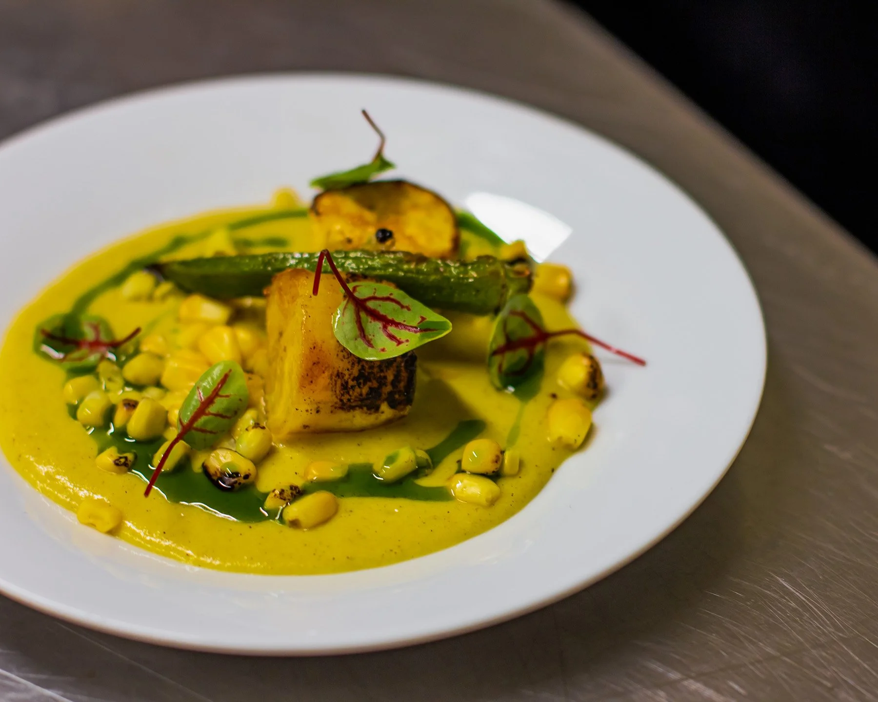 Grilled vegetables with corn, zucchini, and yellow squash, garnished with microgreens and served with a yellow sauce on a white plate.