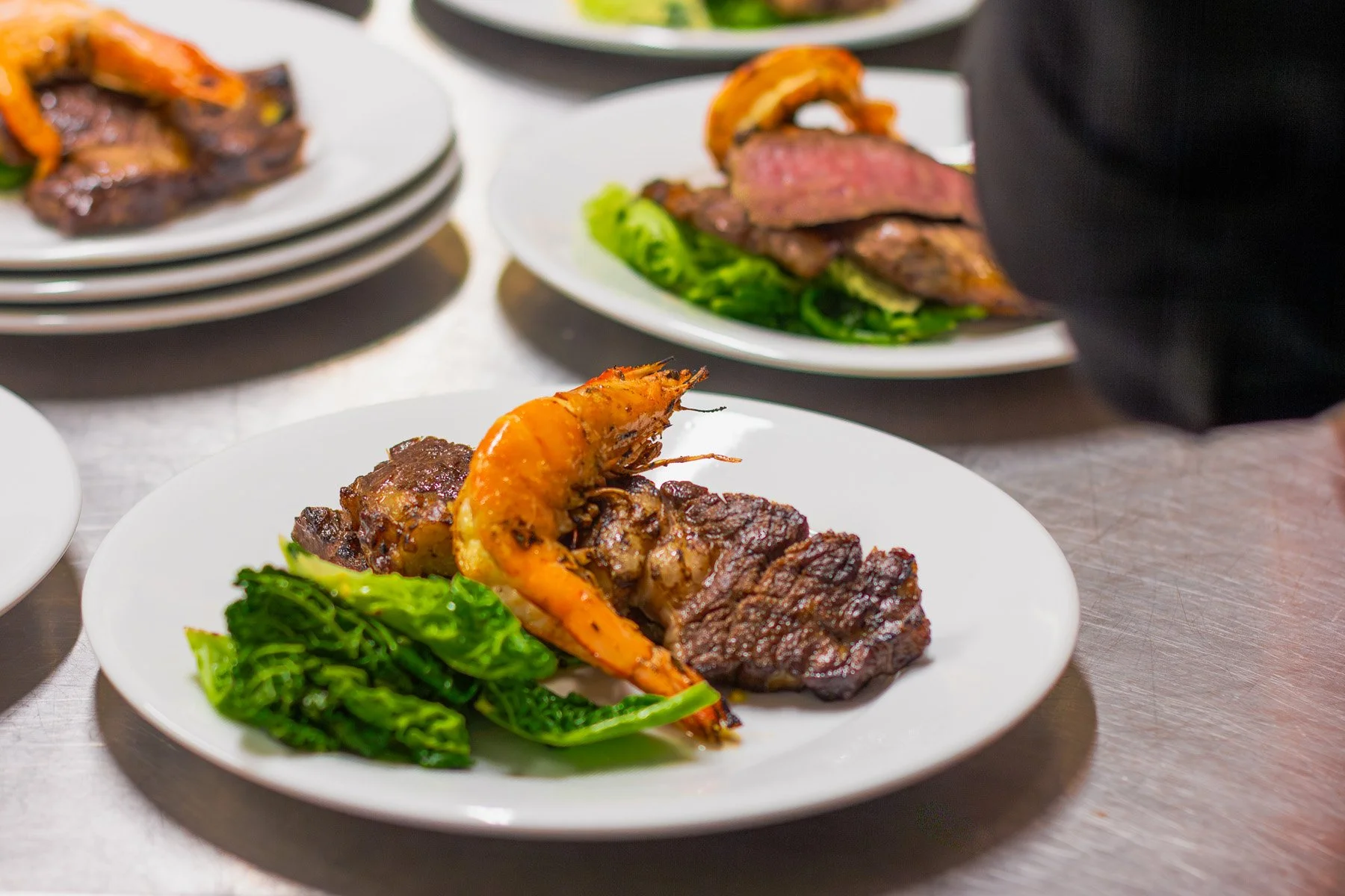 Plate with grilled steak, grilled shrimp, and sautéed greens on a white plate, with other plates of similar food in the background.