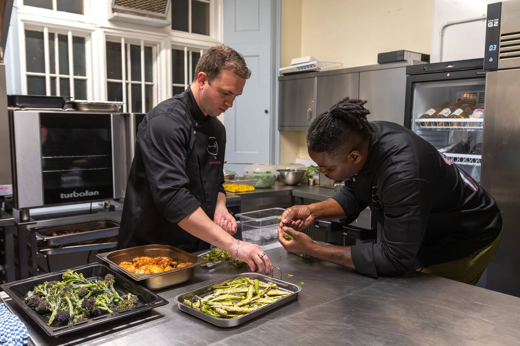 Two chefs preparing food in a commercial kitchen, with trays of broccoli, green beans, and cooked chicken on the counter.