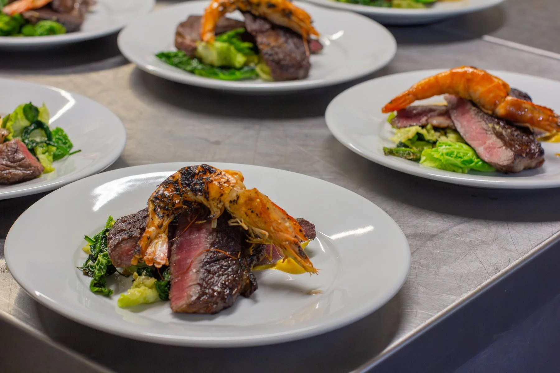 Plates of cooked steak topped with shrimp, served with greens and vegetables on a stainless steel countertop.