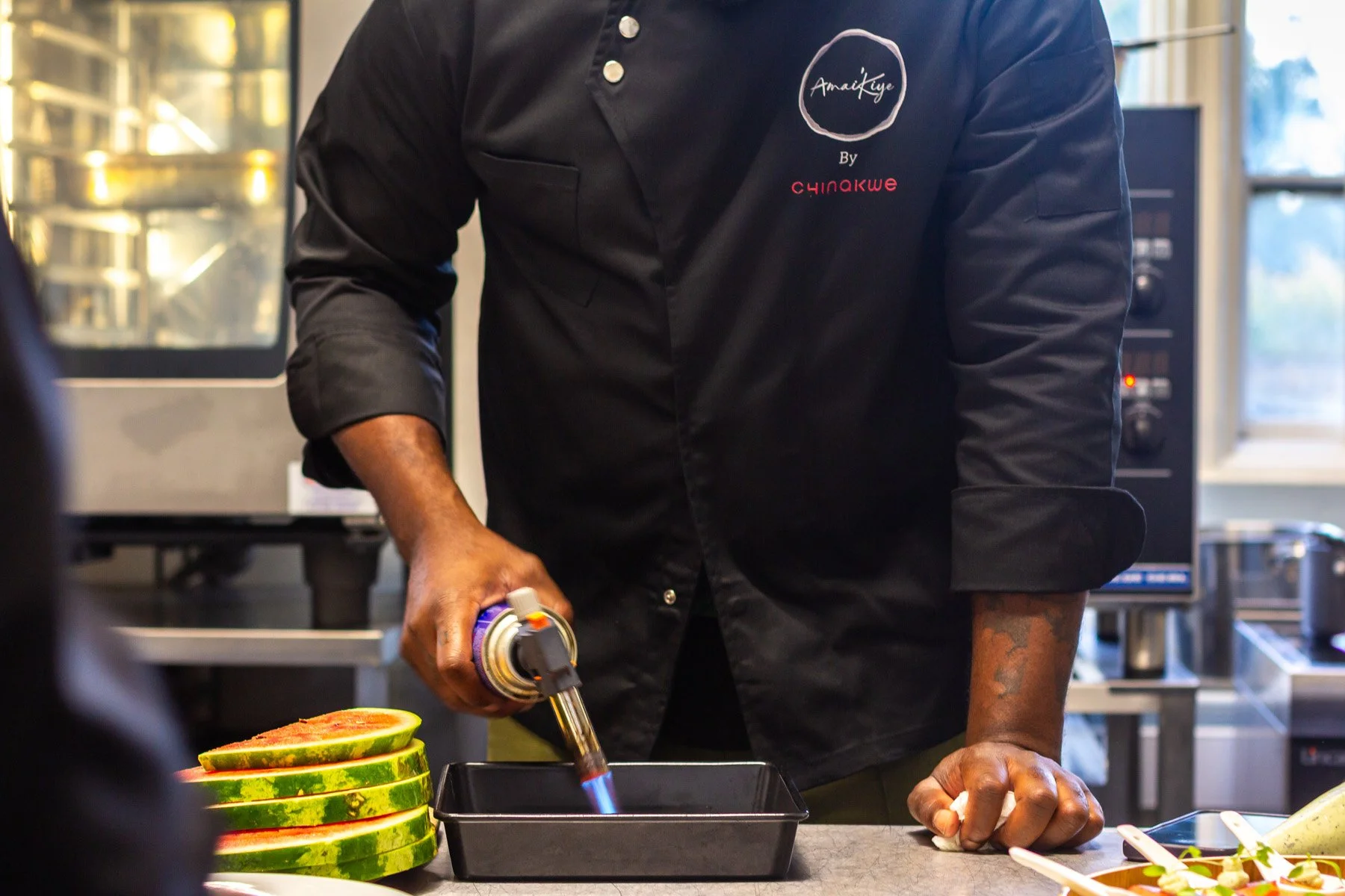 A chef in a black uniform using a culinary torch on a small black tray, with sliced watermelon on the side in a kitchen setting.