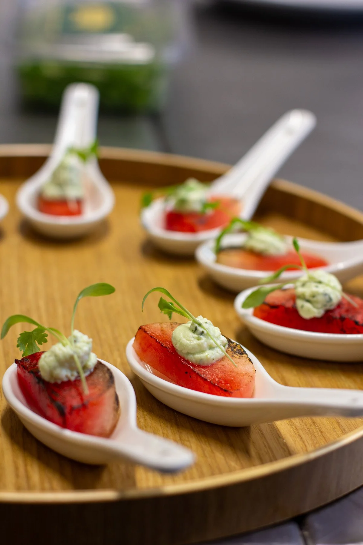 Appetizers served on small white spoons with grilled tomato slices, green herb mousse, and microgreens, arranged on a round wooden tray.