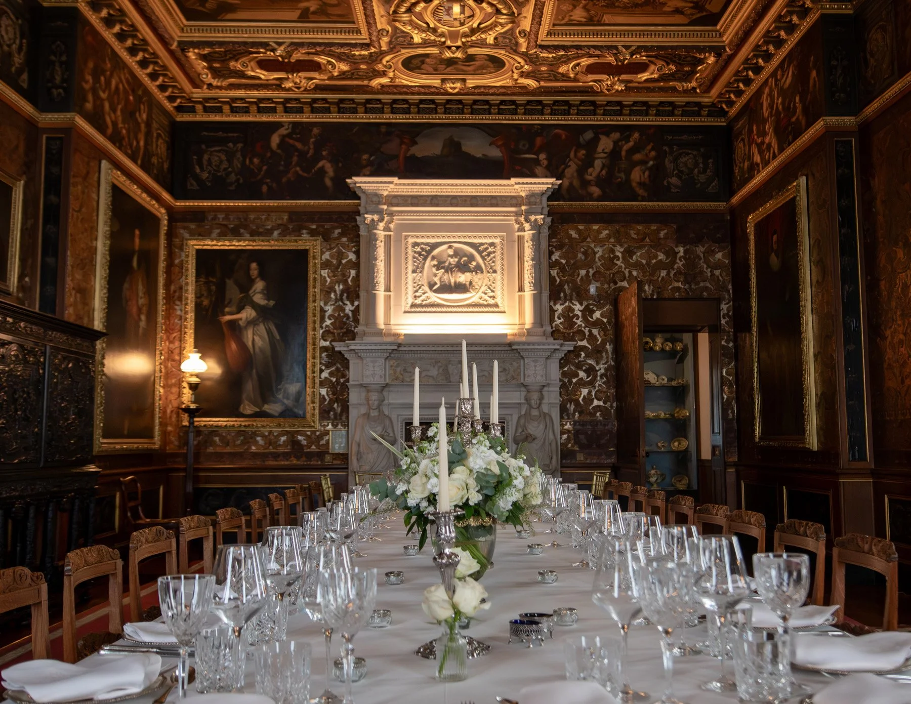 Elegant dining room with a long table set with white tablecloth, crystal glassware, and floral centerpiece, ornate wood-paneled walls, large paintings, and a decorative fireplace with statues.