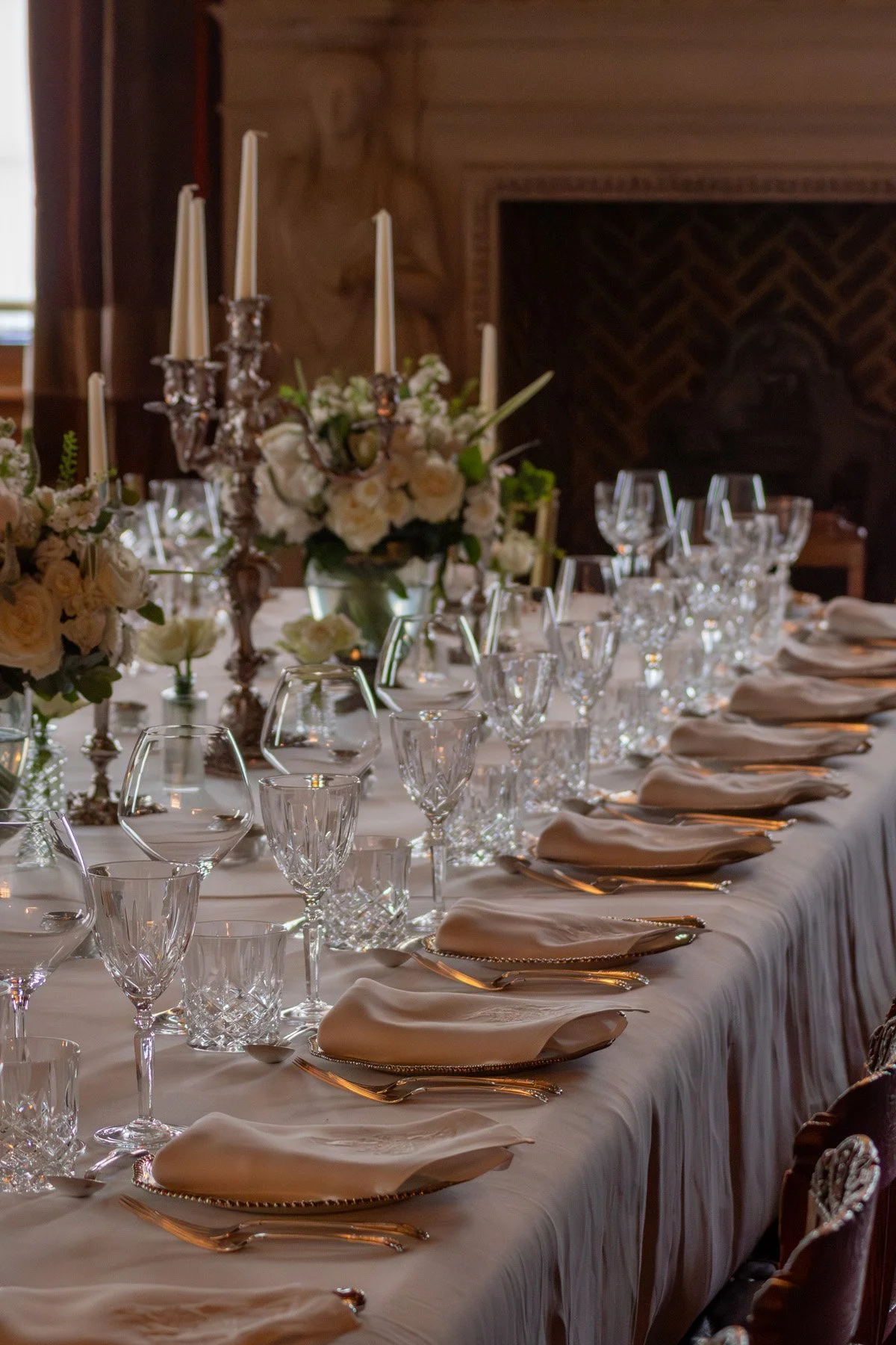 Elegant banquet table set with white tablecloth, crystal glassware, gold utensils, cream napkins, and floral centerpieces with white flowers and candles in a wood-paneled room.