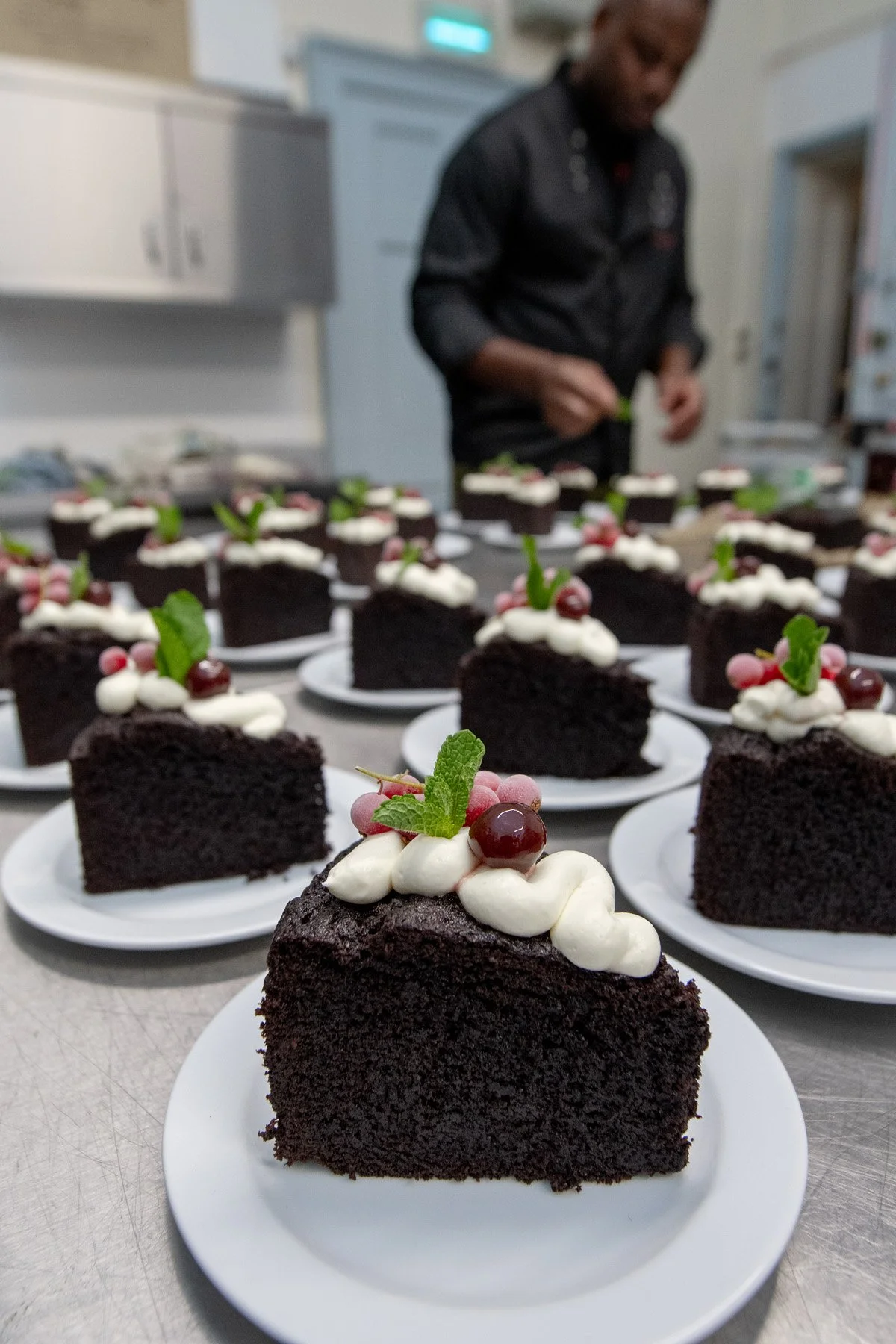 Several slices of chocolate cake topped with white icing, cherries, and mint leaves on white plates, with a person in a black outfit decorating cakes in the background.