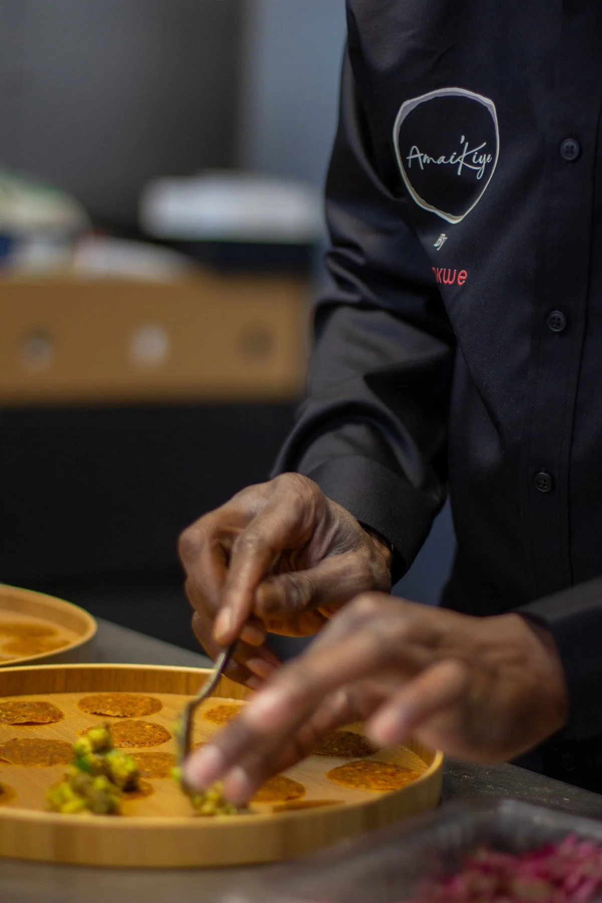 A chef in a black uniform is preparing food, using a small fork to handle a dish with small round items on a bamboo tray.