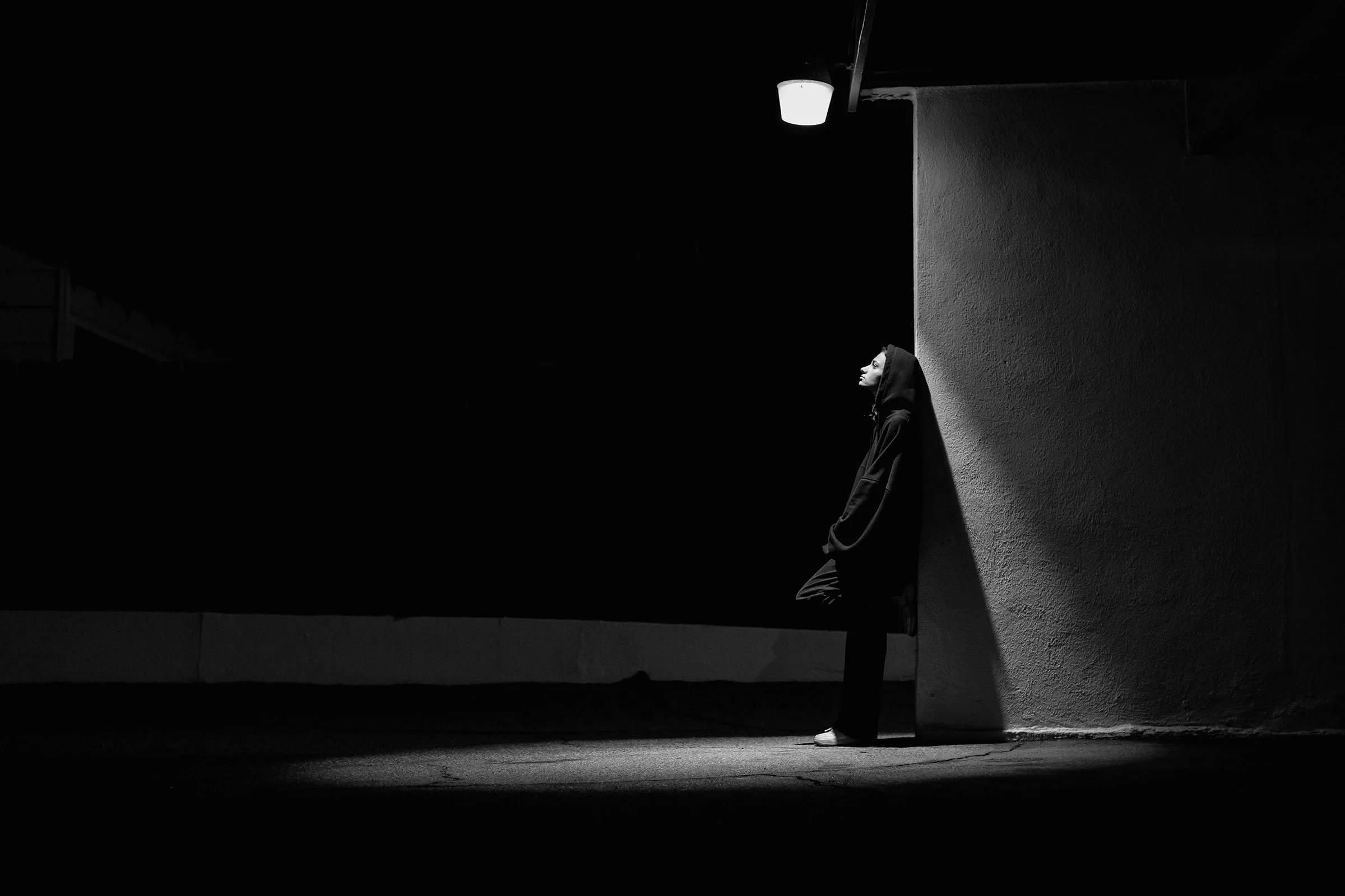 Black and white photo of a person standing against a wall in an urban setting, wearing a hoodie with a shadow cast on their face, illuminated by a streetlamp.