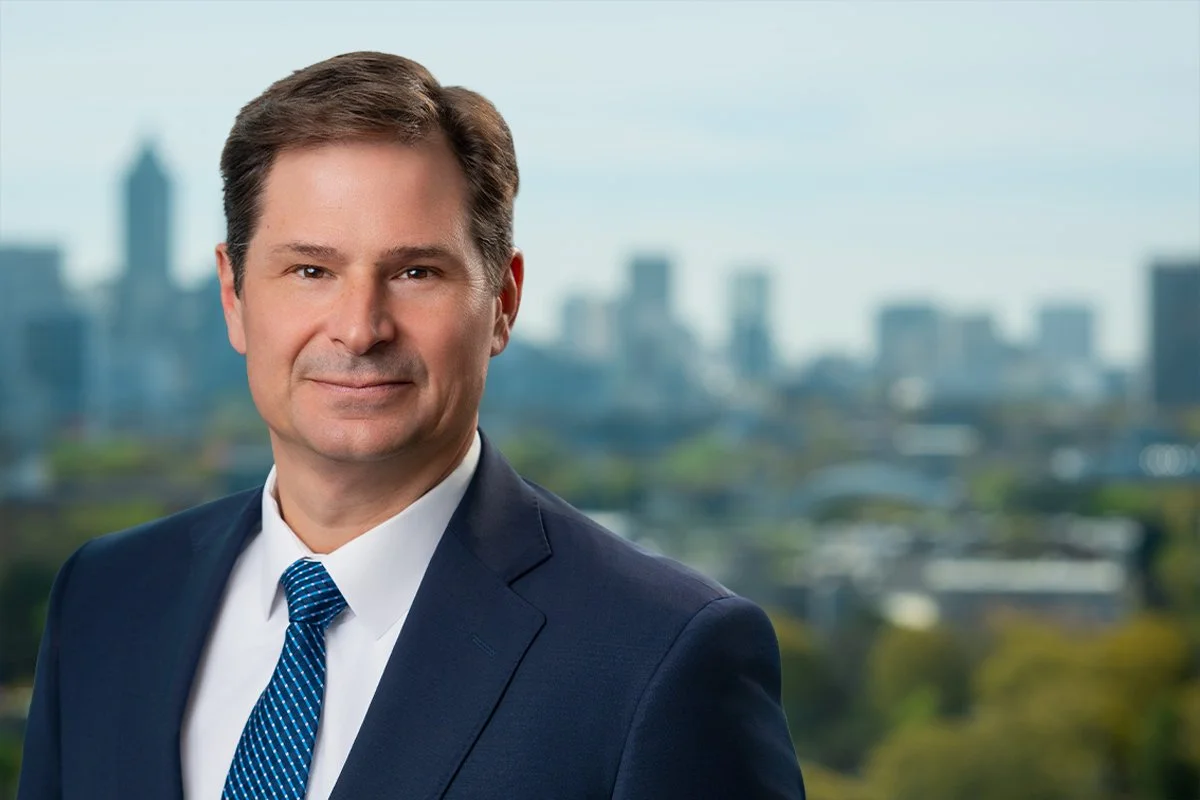 A professional man in a dark suit and blue tie standing outdoors with a city skyline and trees in the background.