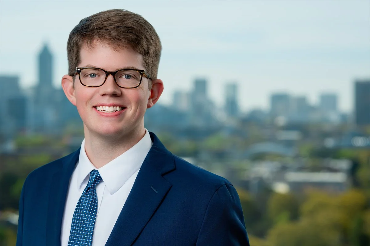 Portrait of a young man wearing glasses, a navy suit, white shirt, and patterned tie, smiling with a city skyline in the background.