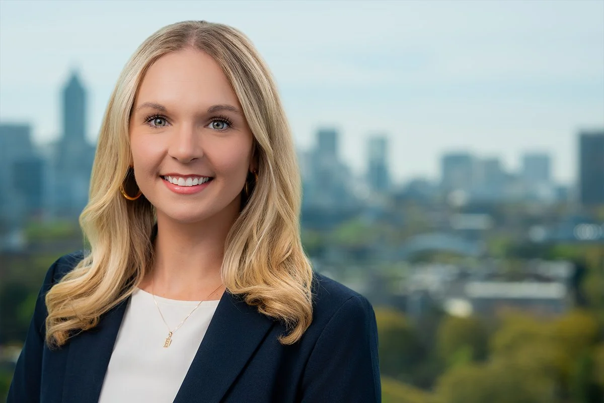 Professional woman with blonde hair smiling, wearing a black blazer and white blouse, against a city skyline background.