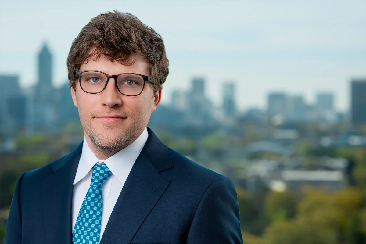 A professionally dressed man wearing glasses, a dark suit, white shirt, and a patterned blue tie, standing outdoors with a blurred city skyline in the background.
