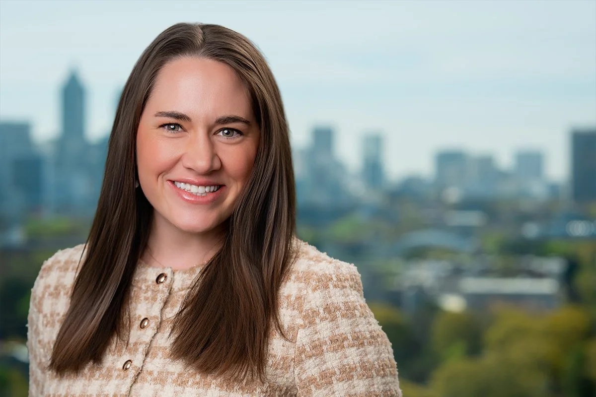 A woman with long brown hair smiling, wearing a dark green blouse with a bow tie, standing with a city skyline in the background.