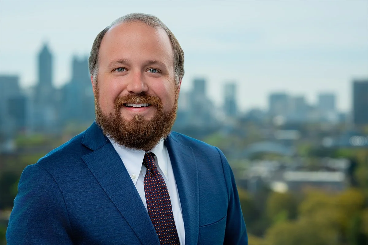 A man with a beard and mustache, wearing a black suit, white shirt, and a red patterned tie, smiling with a city skyline in the background.