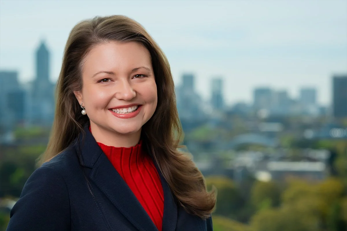 Smiling woman with long brown hair wearing a black blazer and red top, city skyline in the background.