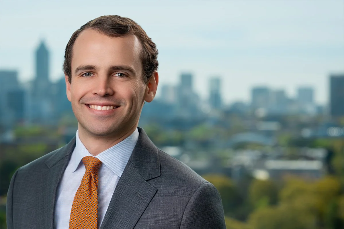 A smiling man in a suit and tie standing outdoors with a city skyline in the background.