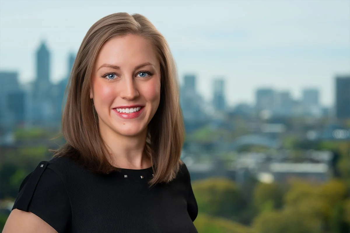 A young woman with long brown hair and blue eyes smiling, with a city skyline in the background.
