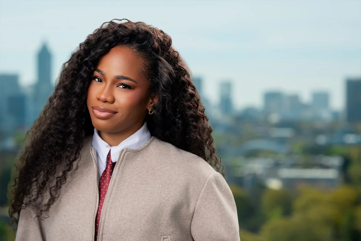 A woman with long curly hair wearing business attire, standing outdoors with a city skyline in the background.