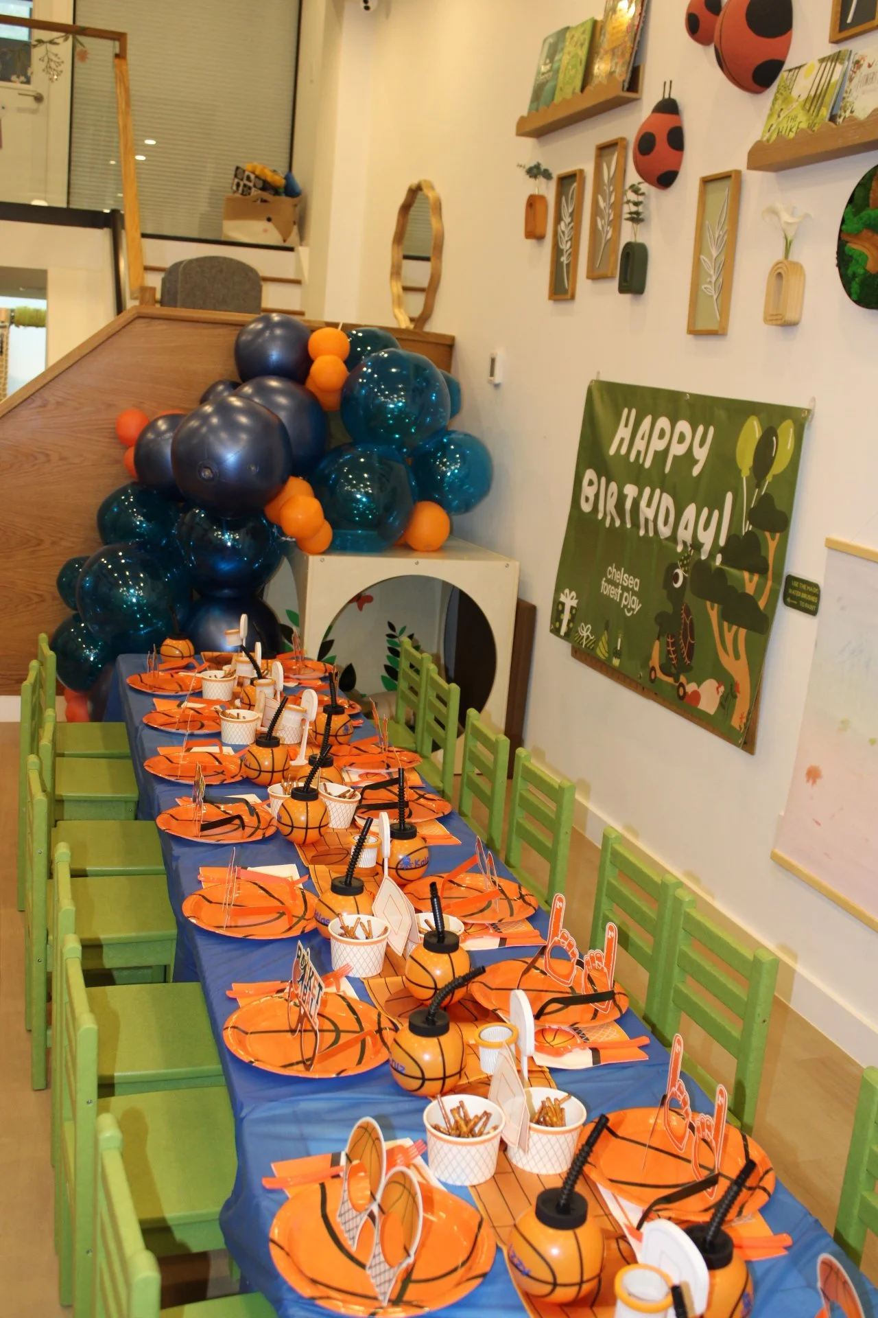 Party decorations with a basketball theme featuring a long table set with orange and black basketball-themed plates, cups, and straws, surrounded by green chairs. The wall is decorated with blue and orange balloons, ladybug and ladybird wall decor, and a 'Happy Birthday' sign with a woodland theme.