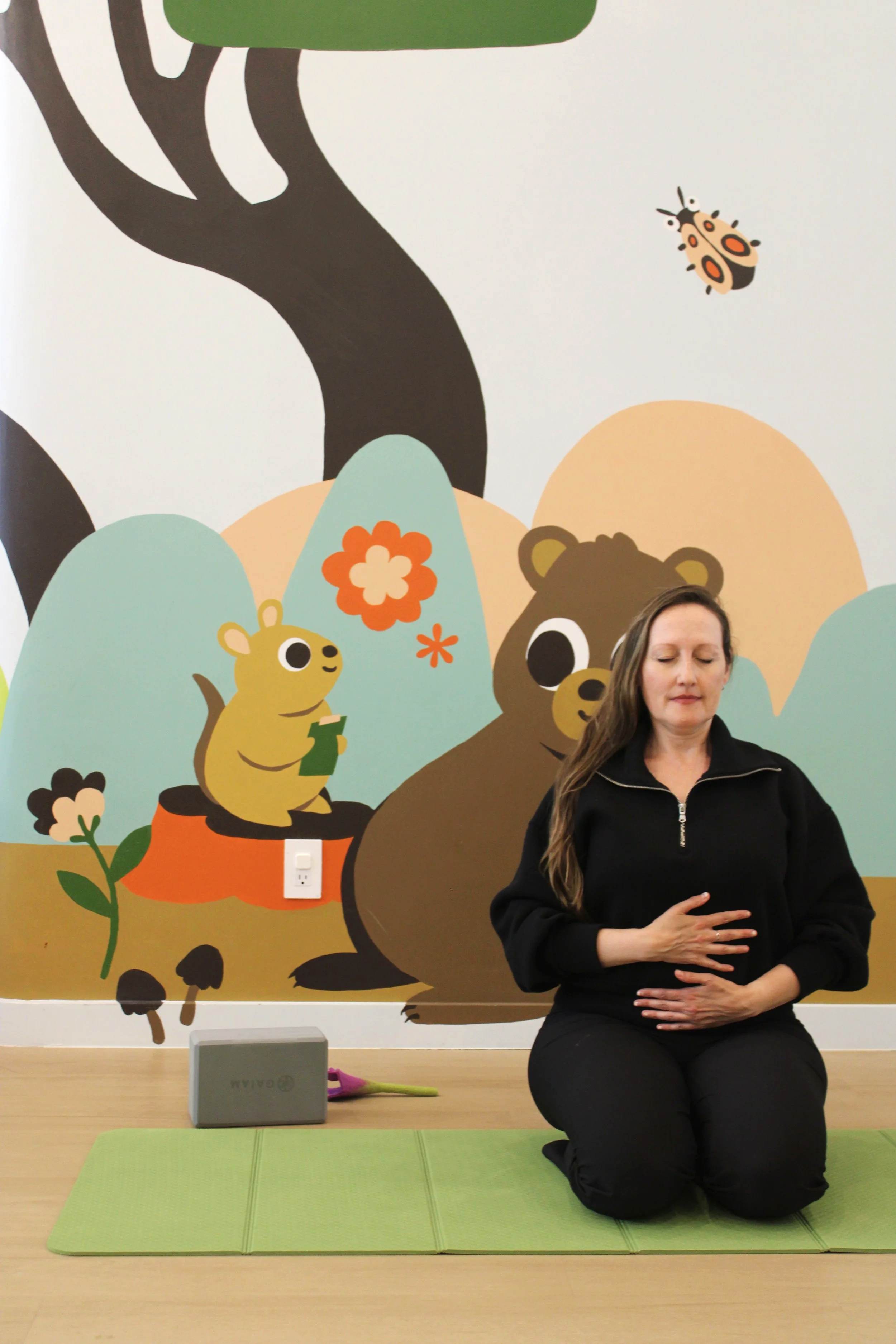 Woman meditating on a green yoga mat, with a colorful cartoon wall mural of animals and trees behind her, and a small gray speaker and notebook on the floor nearby.