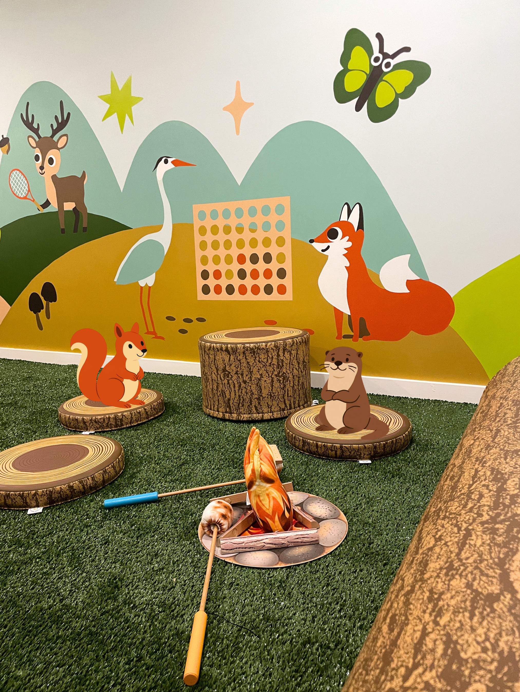 Decorative wall with cartoon animals including a fox, deer, stork, and squirrel, set on a green grass-like surface with faux tree stumps and a wooden log. The wall features illustrated mountains, butterfly, stars, and a connect-four game.
