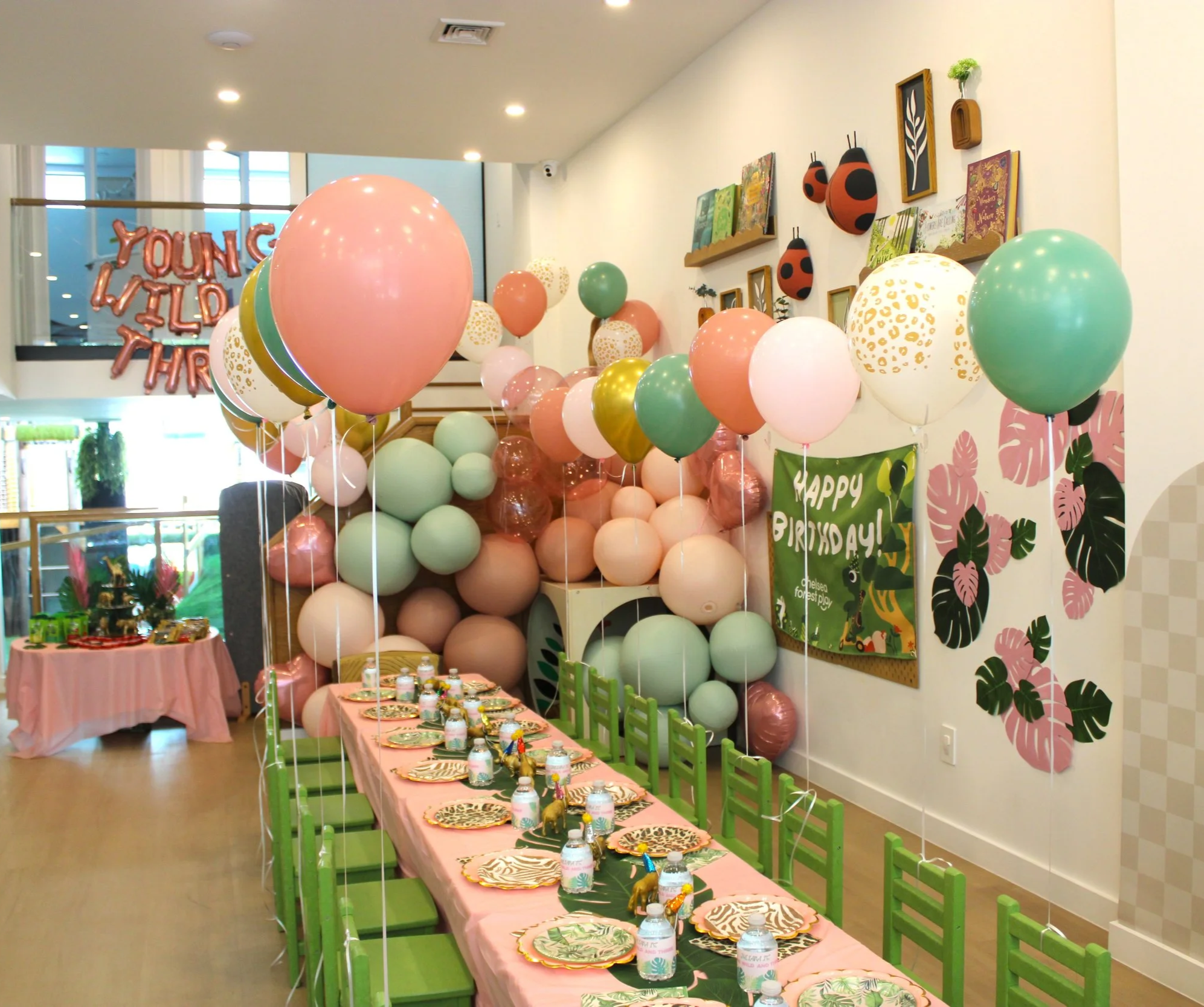 A decorated indoor birthday party setup with a long pink table, green chairs, colorful balloons, and tropical-themed decorations including a "Happy Birthday" banner and tableware.