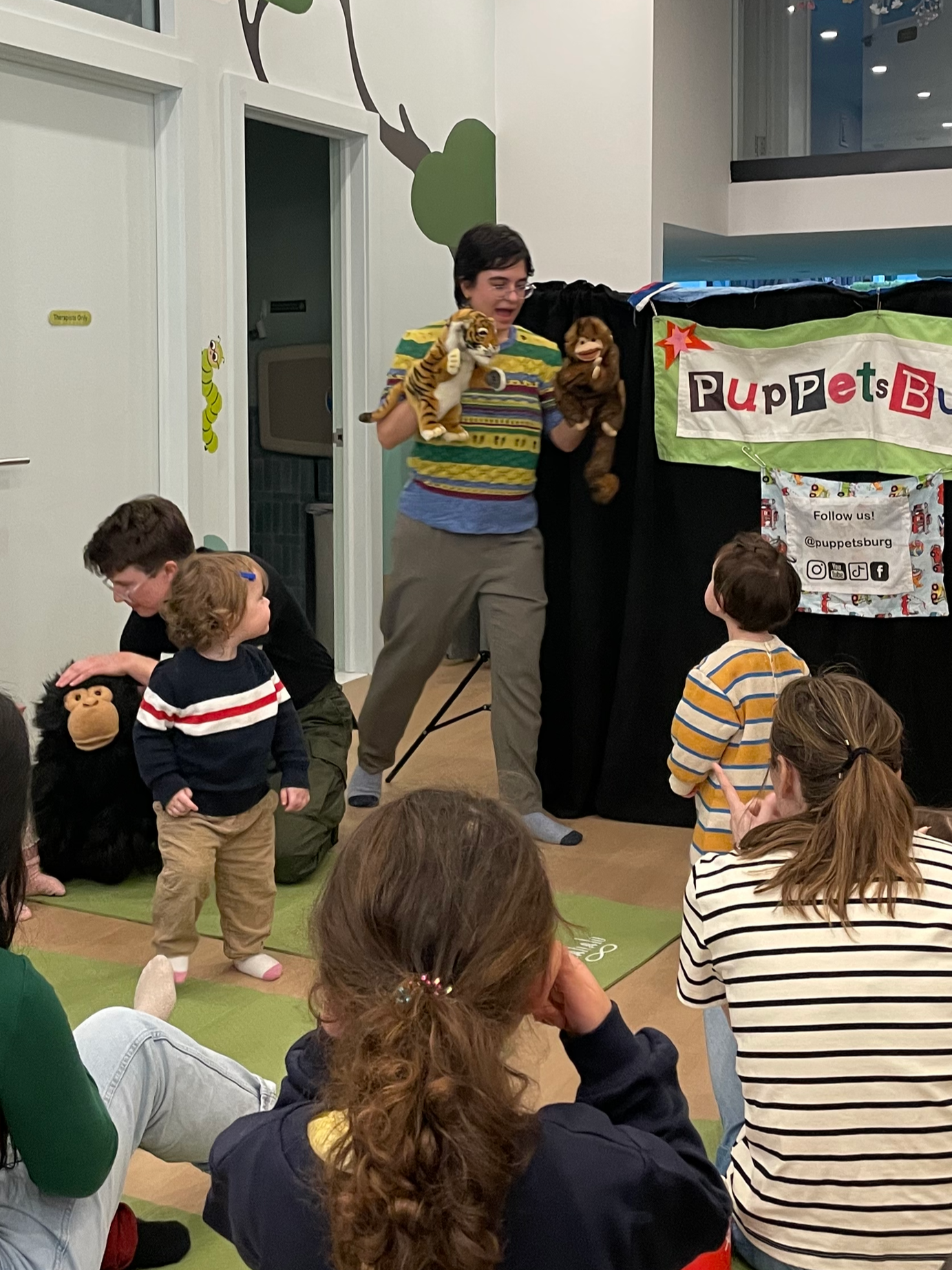 A puppet show with children and adults watching. The performer is holding two puppets, a tiger and a monkey, and is engaging with the audience. The background has a banner that reads 'PuppetsBurg'