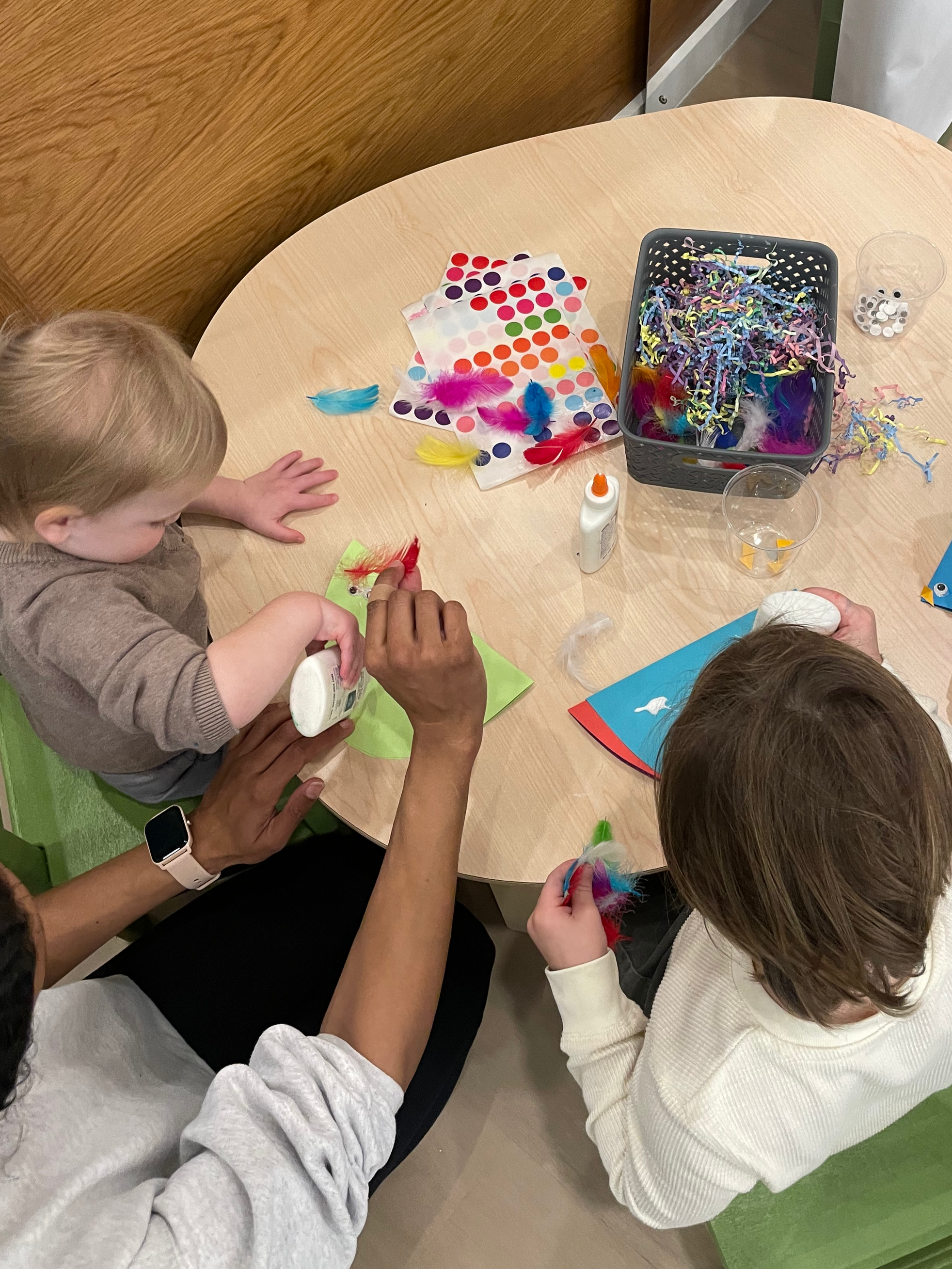 Two children and an adult at a round table engaged in arts and crafts using feathers, glue, and colorful paper, attending an art class at chelsea forest play, an indoor play space.