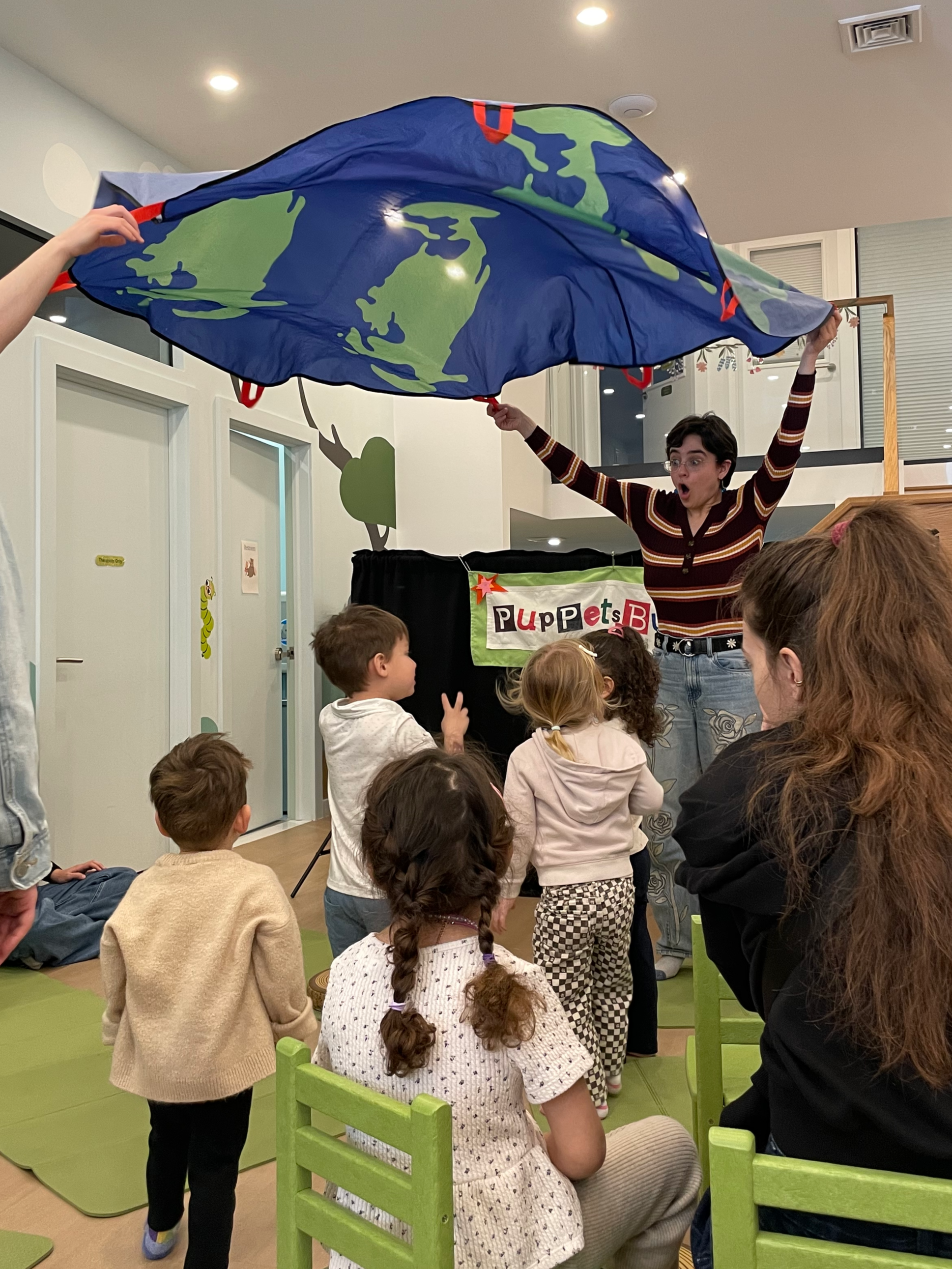 Children and adults watching a performer with a puppet show under a large, hanging Earth-themed parachute