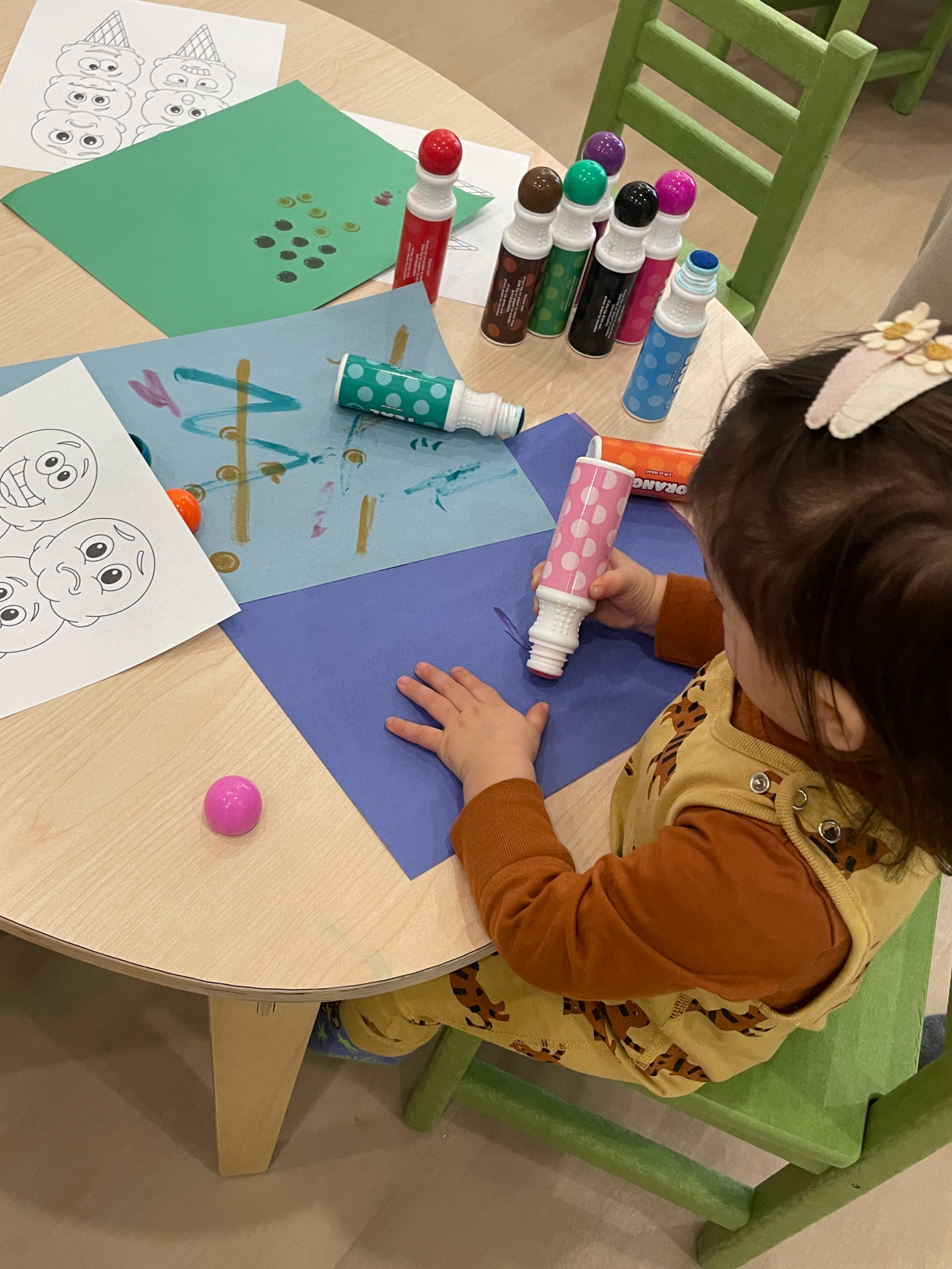 A young child sitting at a wooden table engaged in arts and crafts. They are holding a pink paint tube and applying paint to a blue sheet of paper. The table is scattered with various craft supplies, including bottles of paint, color pages with carto