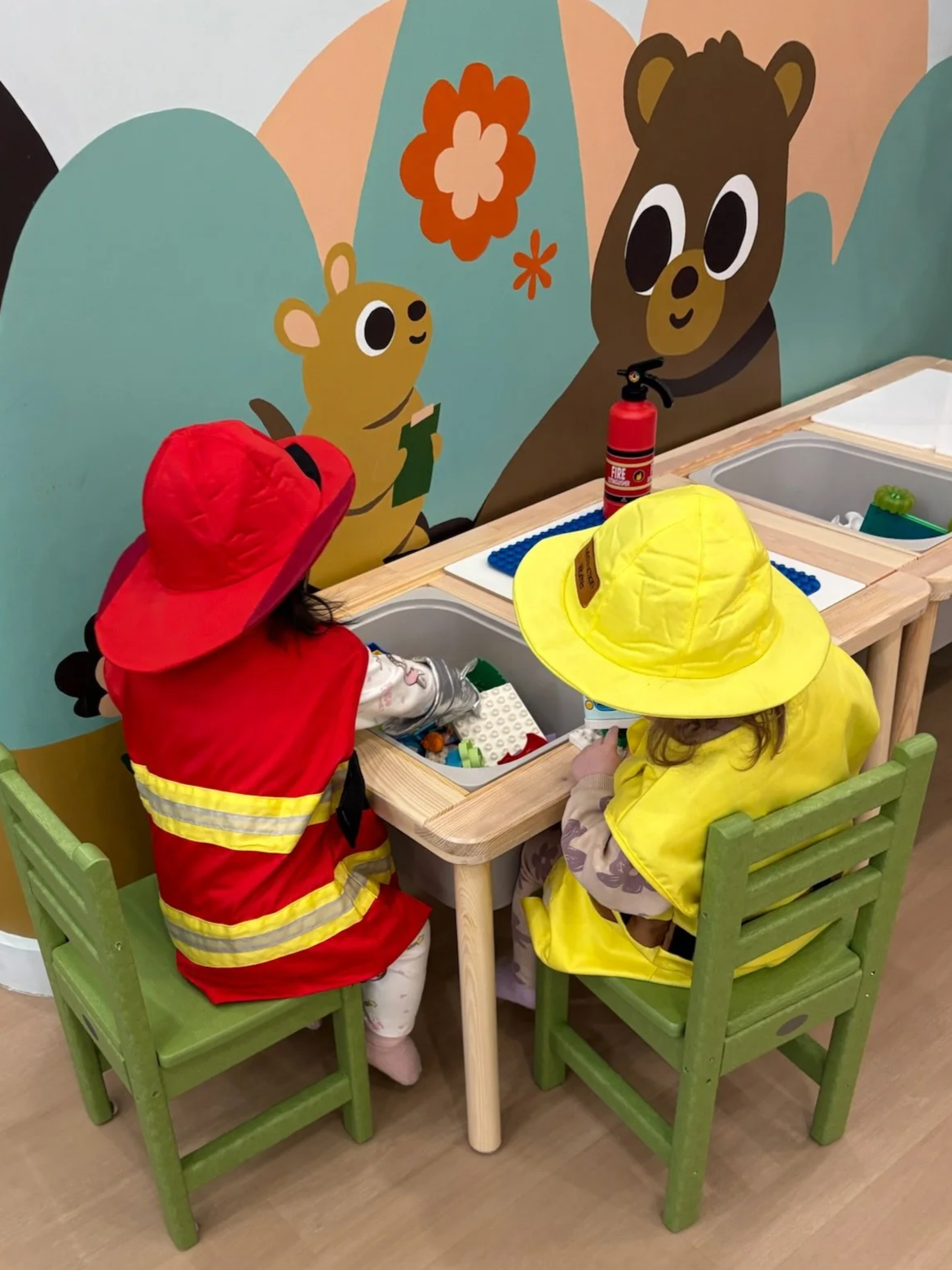 Two children dressed as firefighters playing with toy building blocks at a small table, with a colorful mural of cartoon bears in the background.