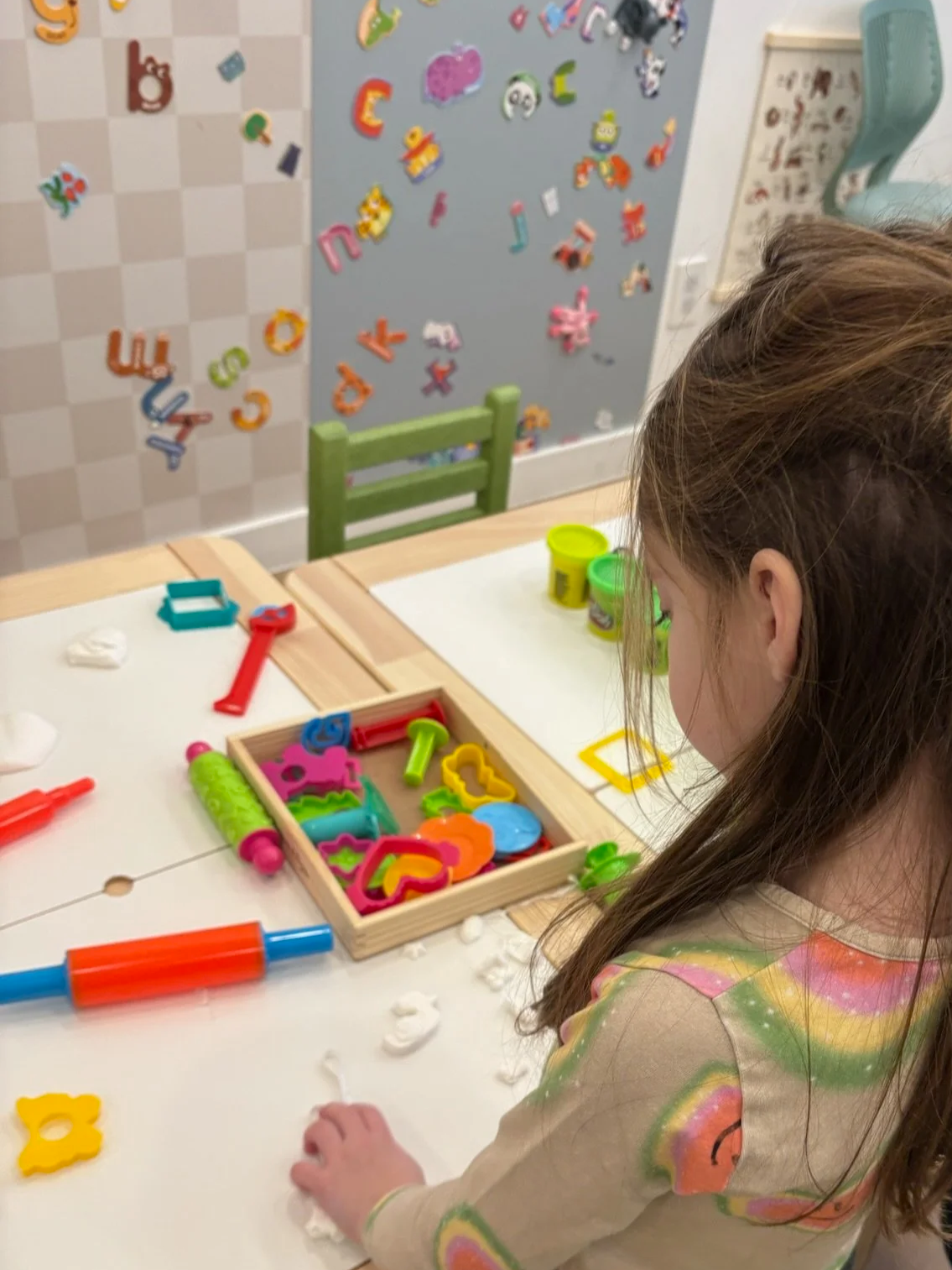 A young girl playing with colorful Play-Doh and cookie cutters at a white table in a playroom with foam alphabet letters on the wall.