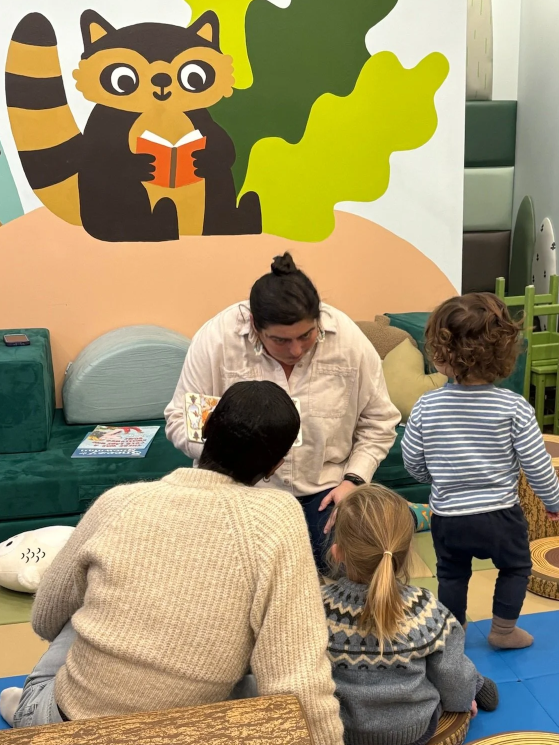 A person with dark hair in a bun reads a story to a group of kids in a colorful room with a raccoon cartoon painted on the wall. Two children sit on the floor, one boy with curly hair and a striped shirt, and a girl with blonde hair in a ponytail, we