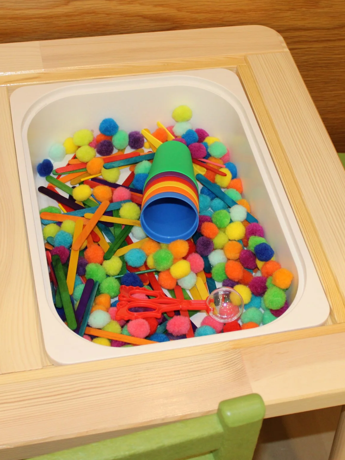 Colorful sensory table filled with multicolored pom-poms, plastic craft sticks, a small plastic scoop, and a stacking cup toy.