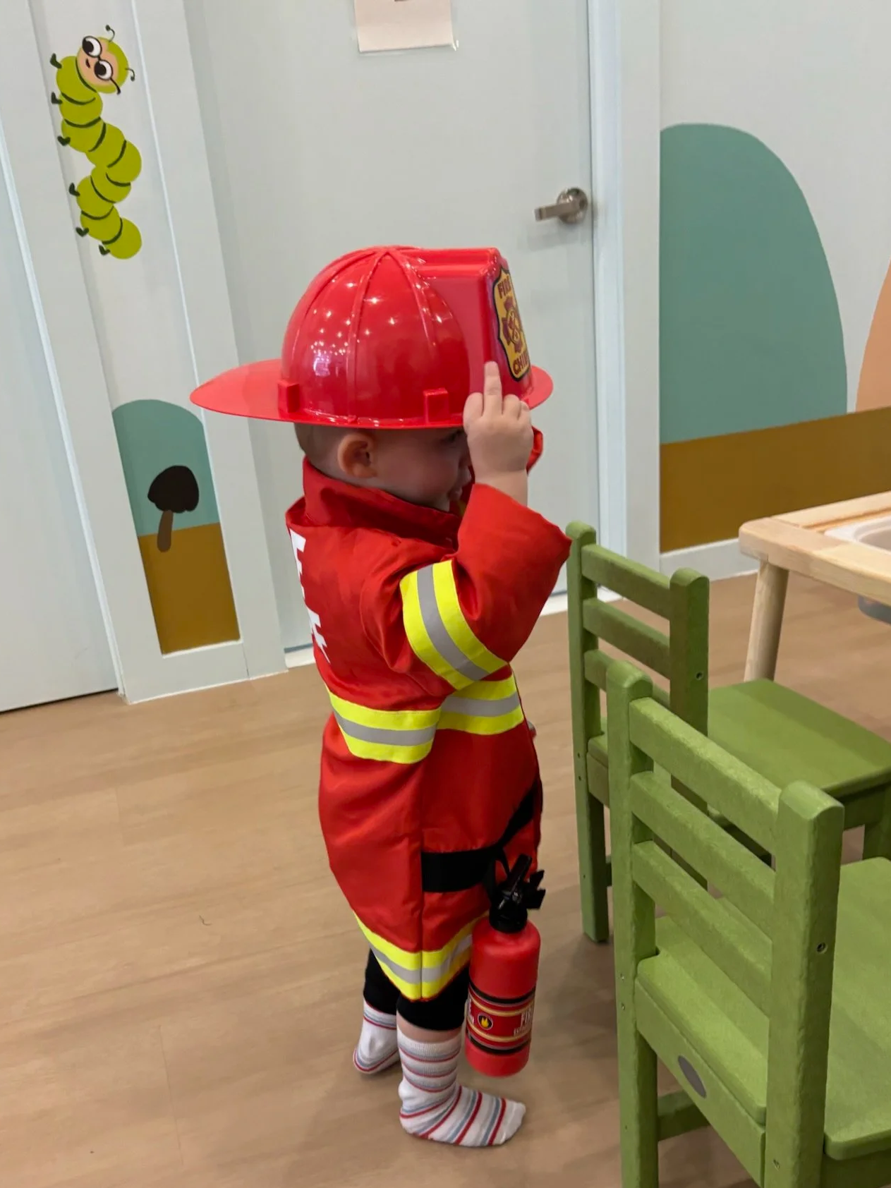 Young child dressed as a firefighter wearing a red helmet and costume, holding a toy fire extinguisher, standing near a green chair in a playroom with wall decals.