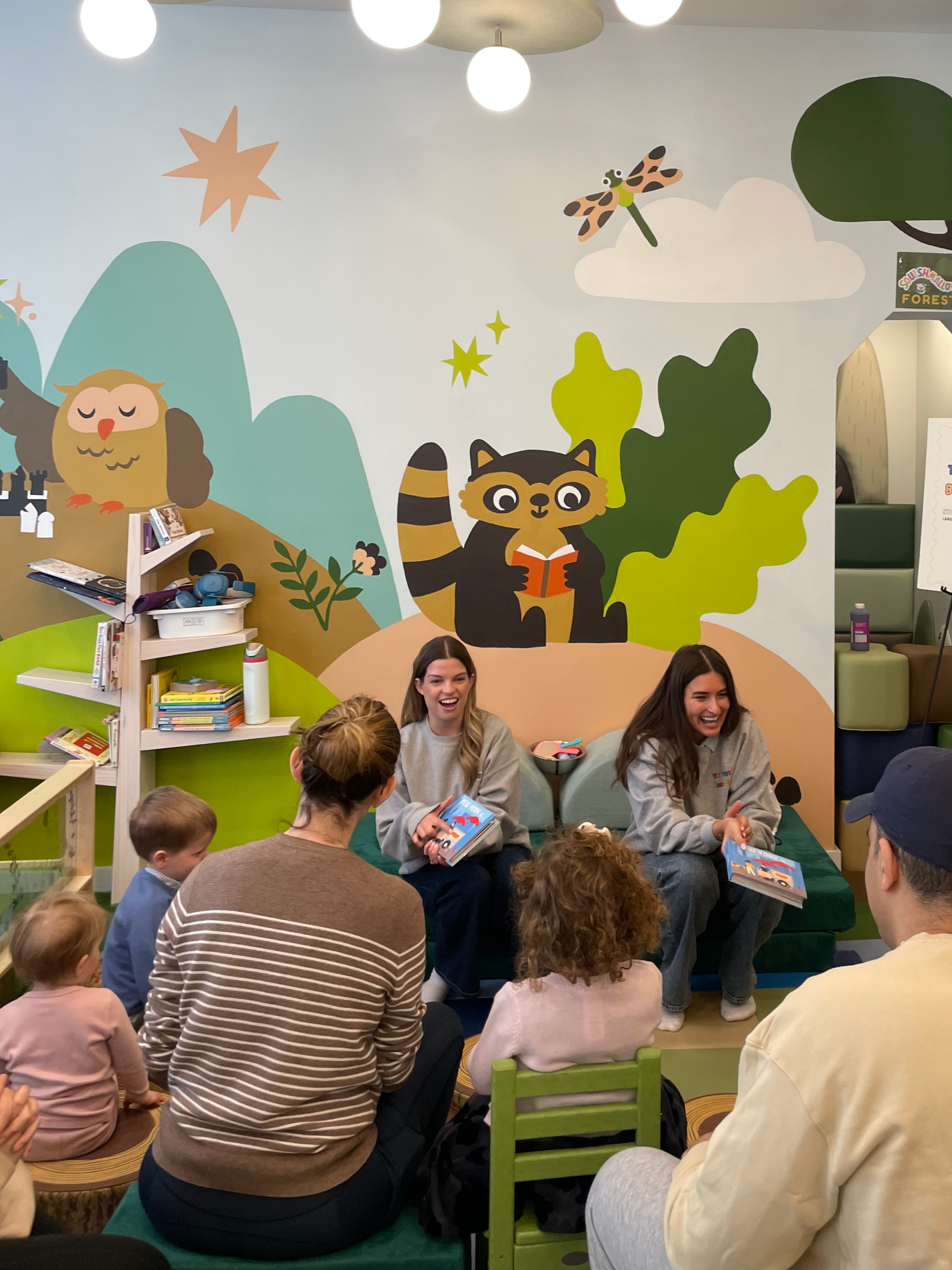 Children and adults sitting on the floor and chairs, listening to two women reading books in a colorful children's library or play area with wall murals of cartoon animals and nature.