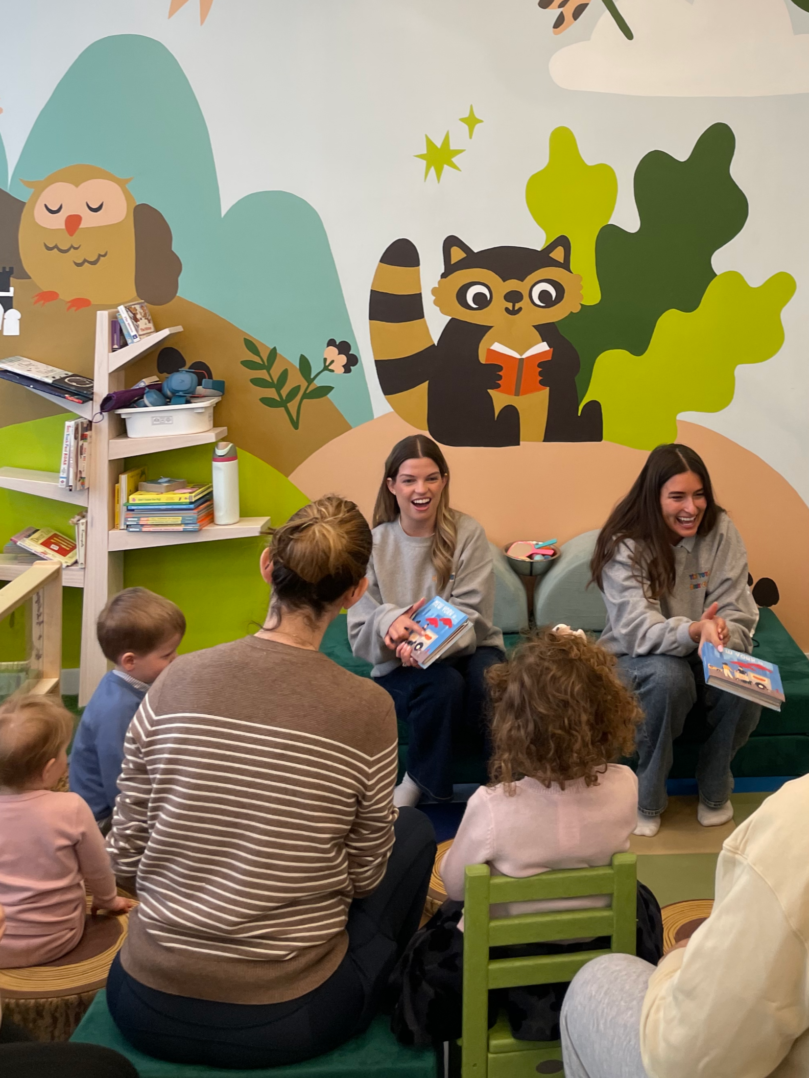 Two women reading children's books to a group of children and adults in a colorful room with a mural of an owl and a raccoon reading books in the background.