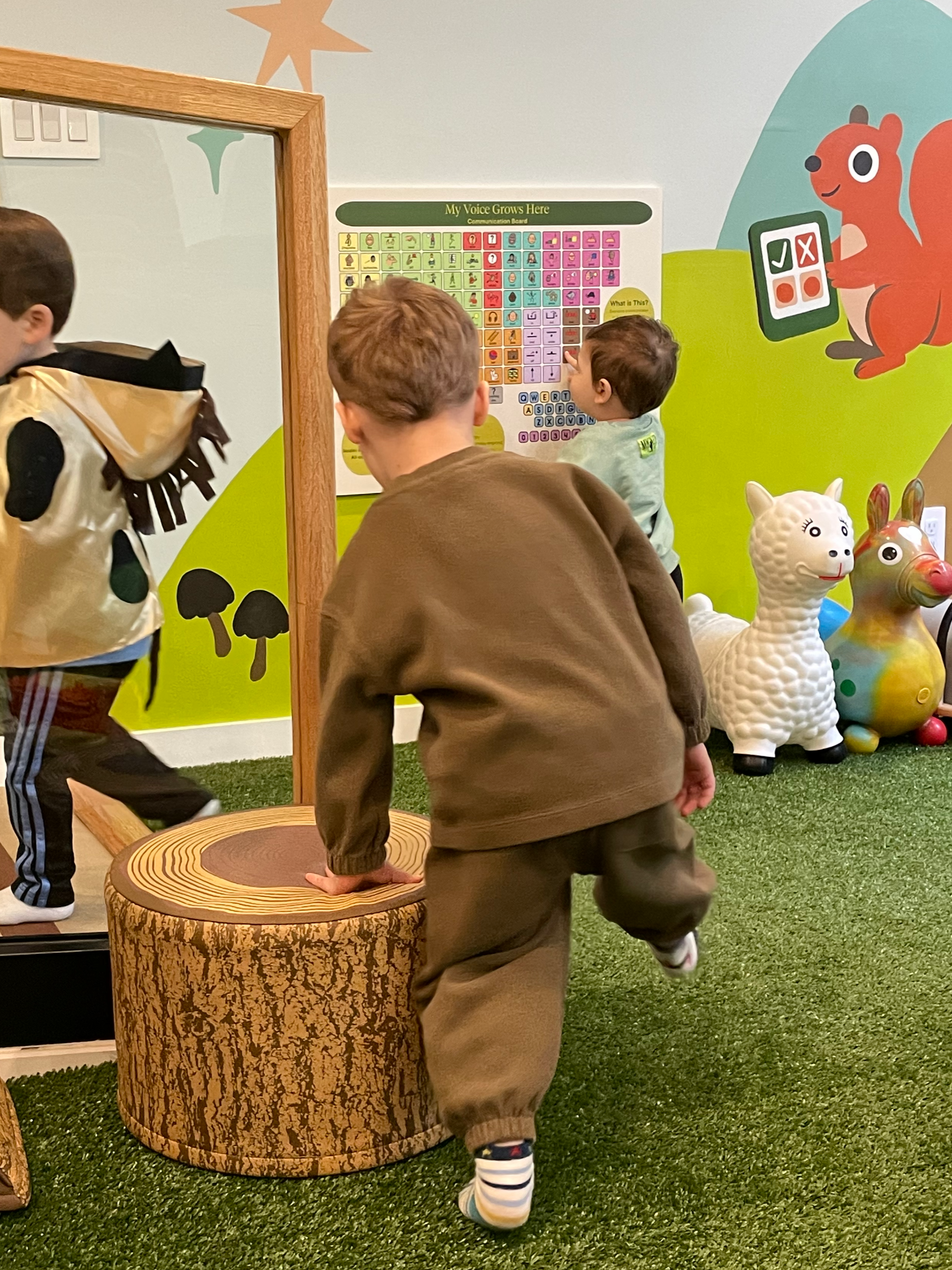 Children playing in a colorful indoor play area with animal toys, a large mirror, and a wall chart titled 'My Voice Grows Here'.