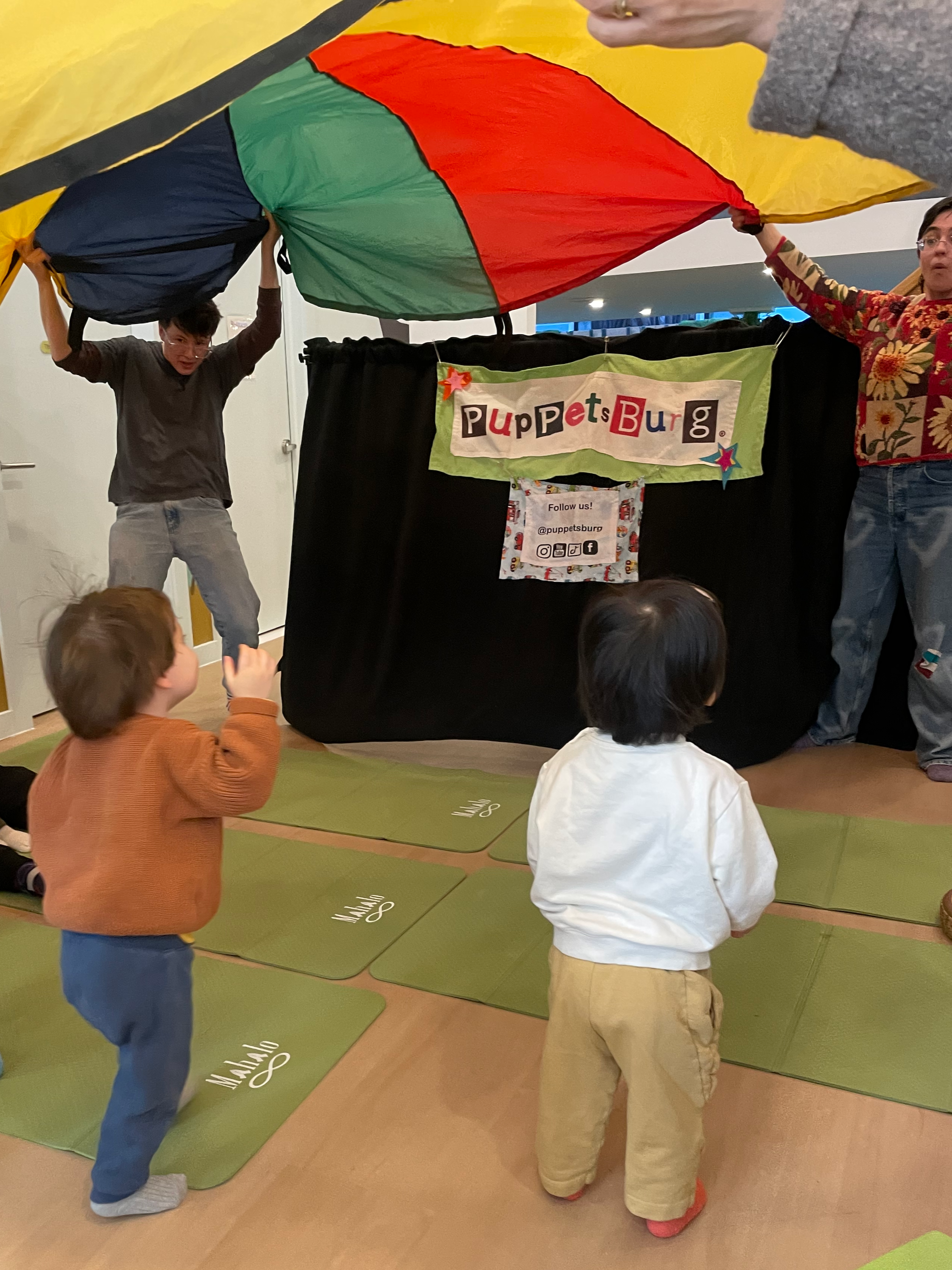 Children watching a puppet show, with a colorful parachute overhead at PuppetsBurg.