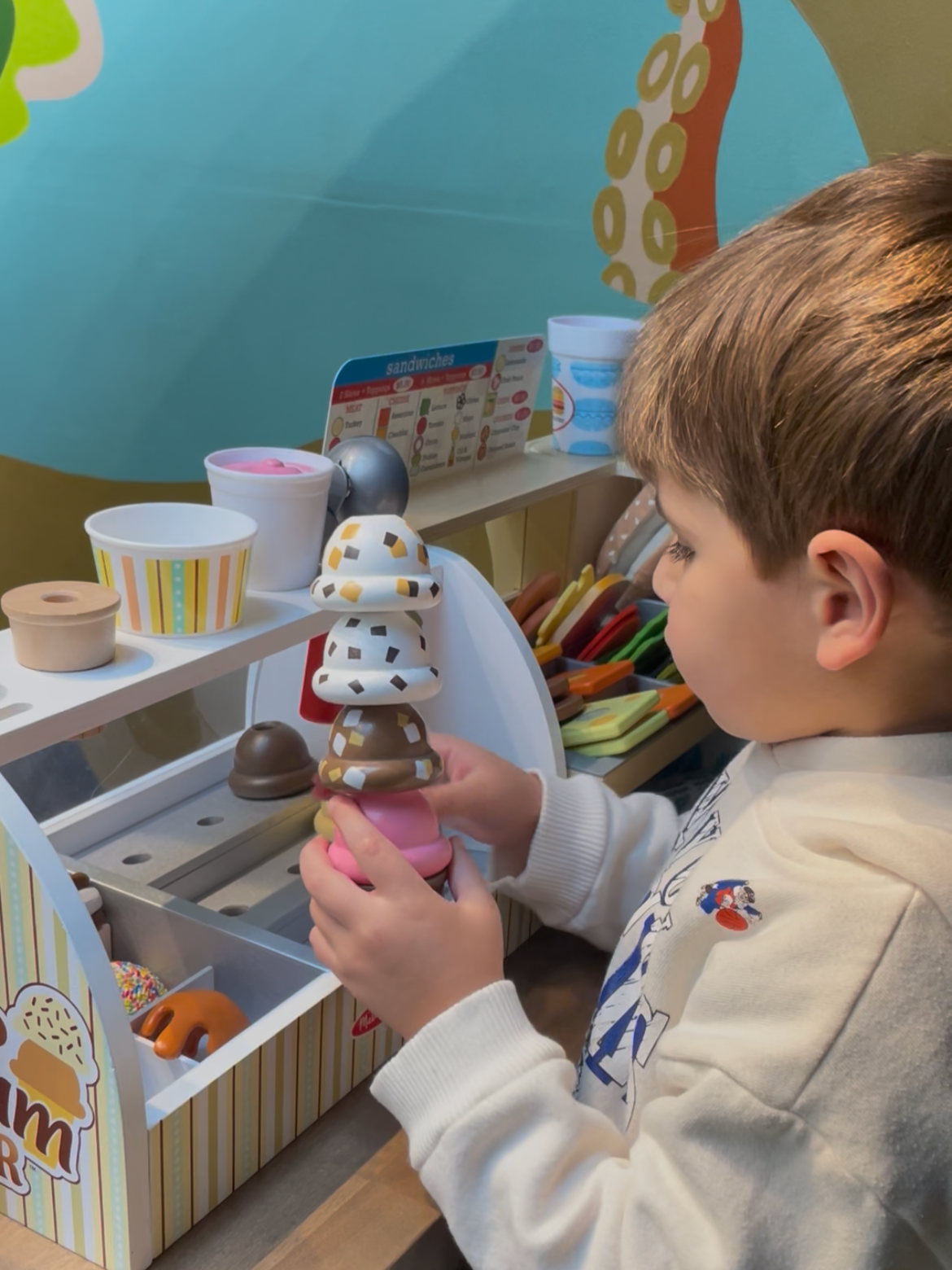 A young boy holding a stack of toy ice cream cones made of plastic in a play ice cream parlor setting.