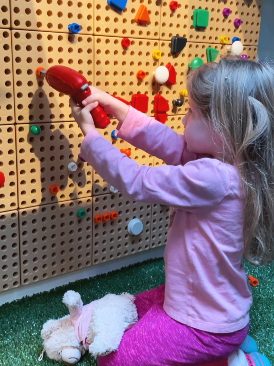 A young girl in pink clothes, with blonde hair, kneeling, playing with a toy hammer in front of a colorful pegboard wall with various shaped and colored pegs, on a green carpet, with a stuffed animal lying next to her.