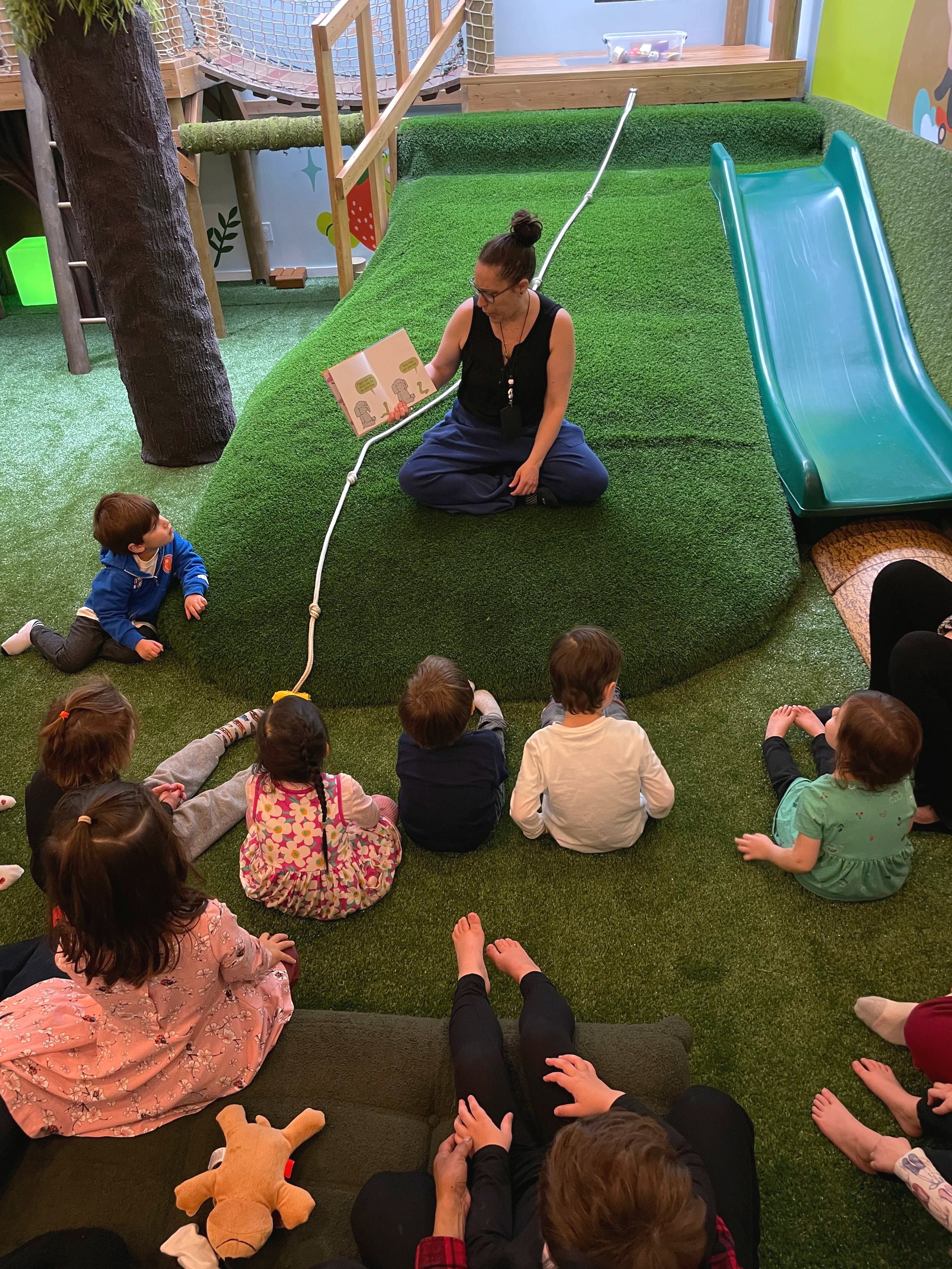 A woman reading a book to a group of children in an indoor play area with green artificial grass, a slide, and a tree stump.