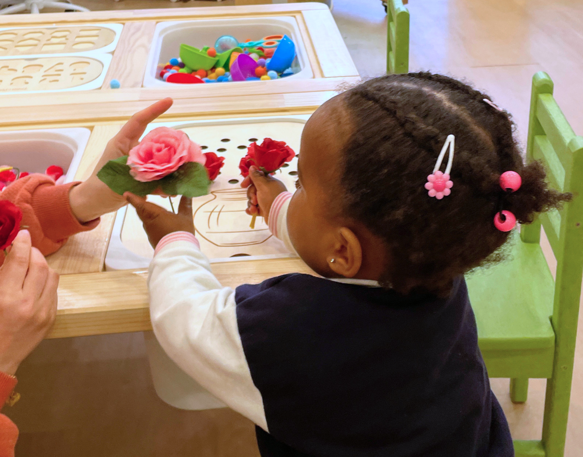 A young girl with braids and pink hair accessories at a table, holding colorful artificial flowers, with a toy ice cream shop setup in front of her.