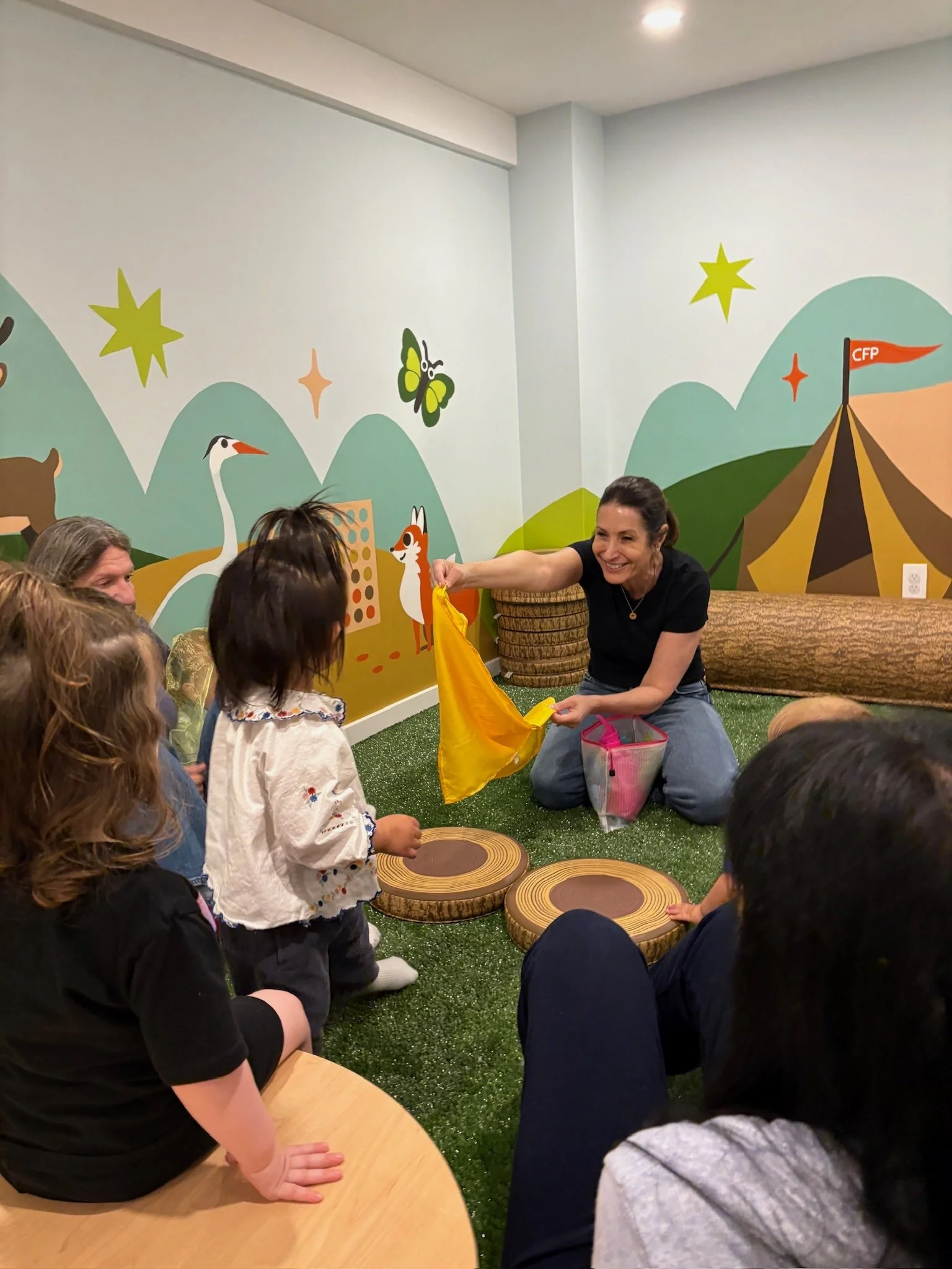 A woman smiling and holding a yellow fabric flag while engaging with children seated on a green carpeted floor with tree trunk-shaped cushions. The background features a colorful mural with mountains, stars, a butterfly, a giraffe, and a tent with a 