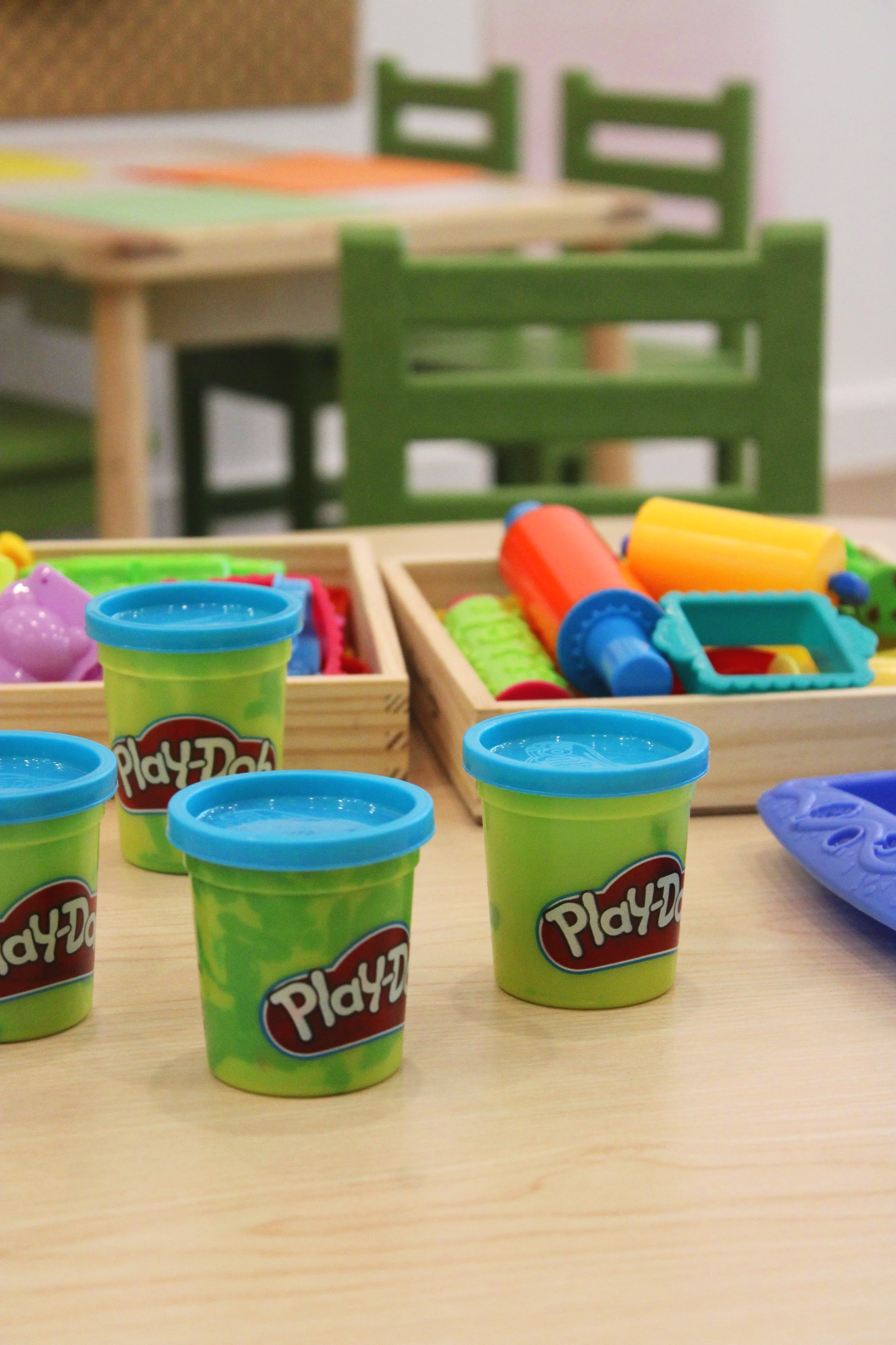 Colorful playdough containers on a table with green and wooden chairs in the background.