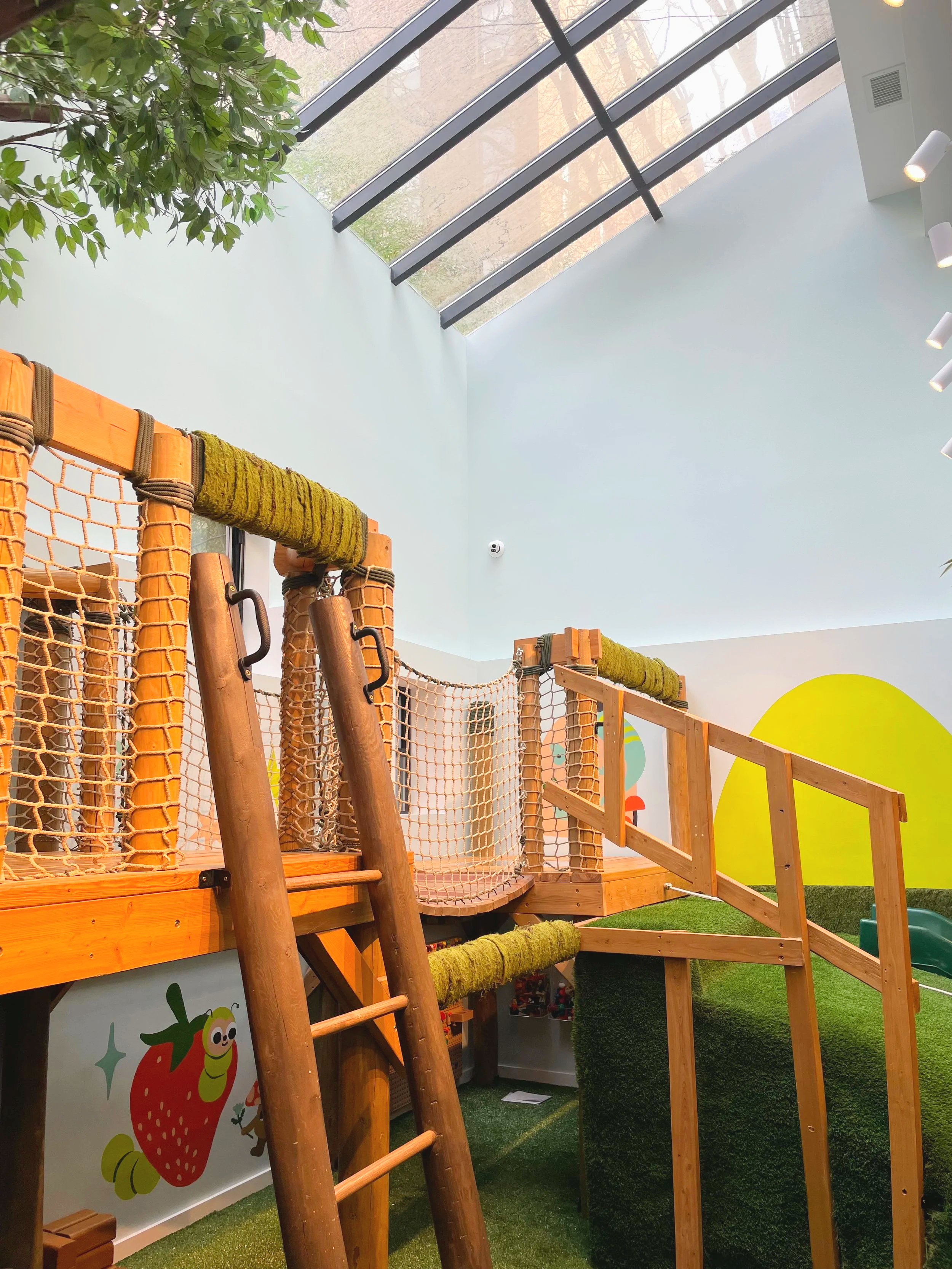 Indoor children's play area with wooden climbing structure, netting, green grass-like flooring, and a large window ceiling allowing natural light.