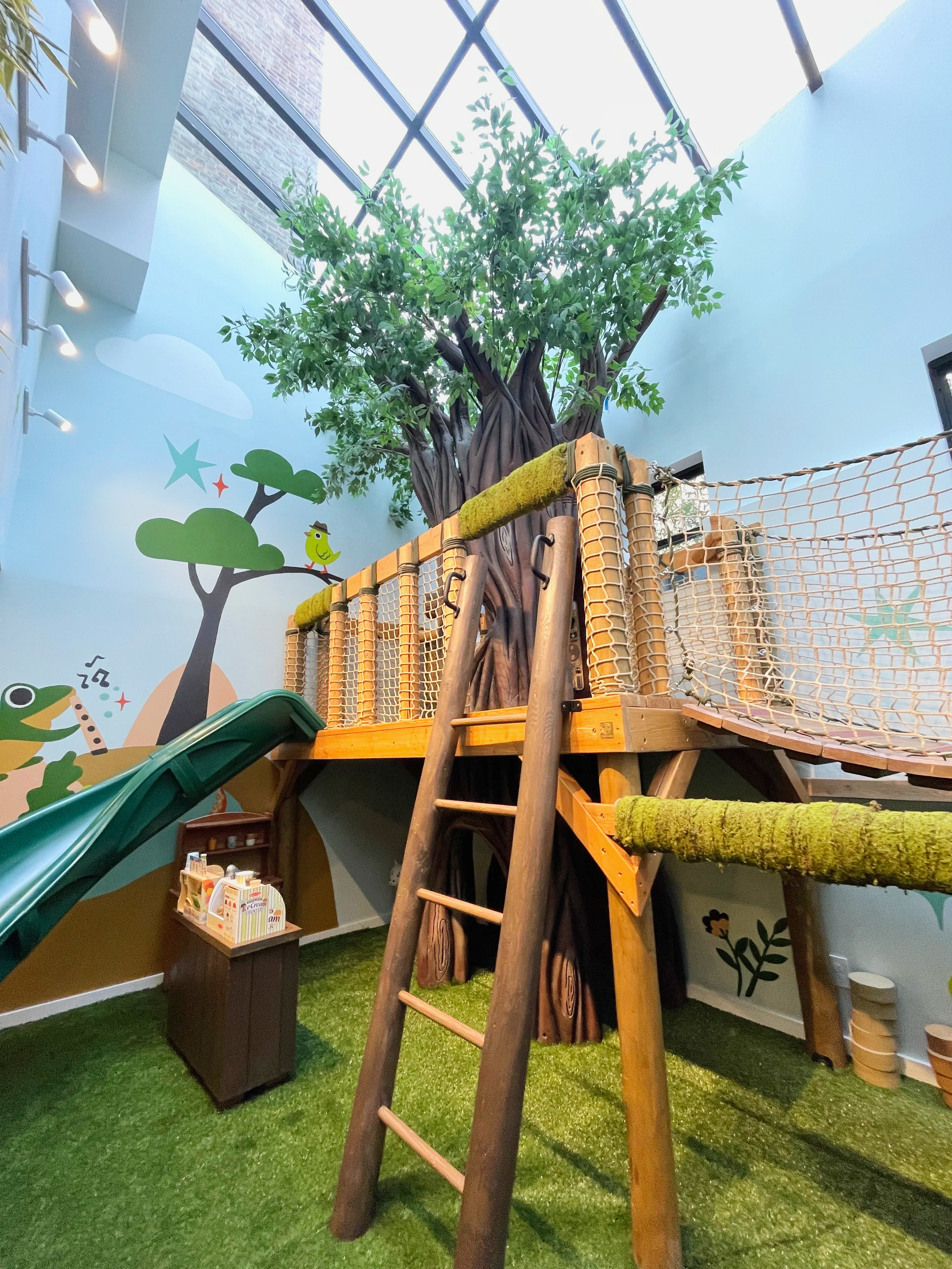 Indoor children's play area with artificial grass, a slide, a treehouse structure, and painted mural featuring cartoon animals and trees.